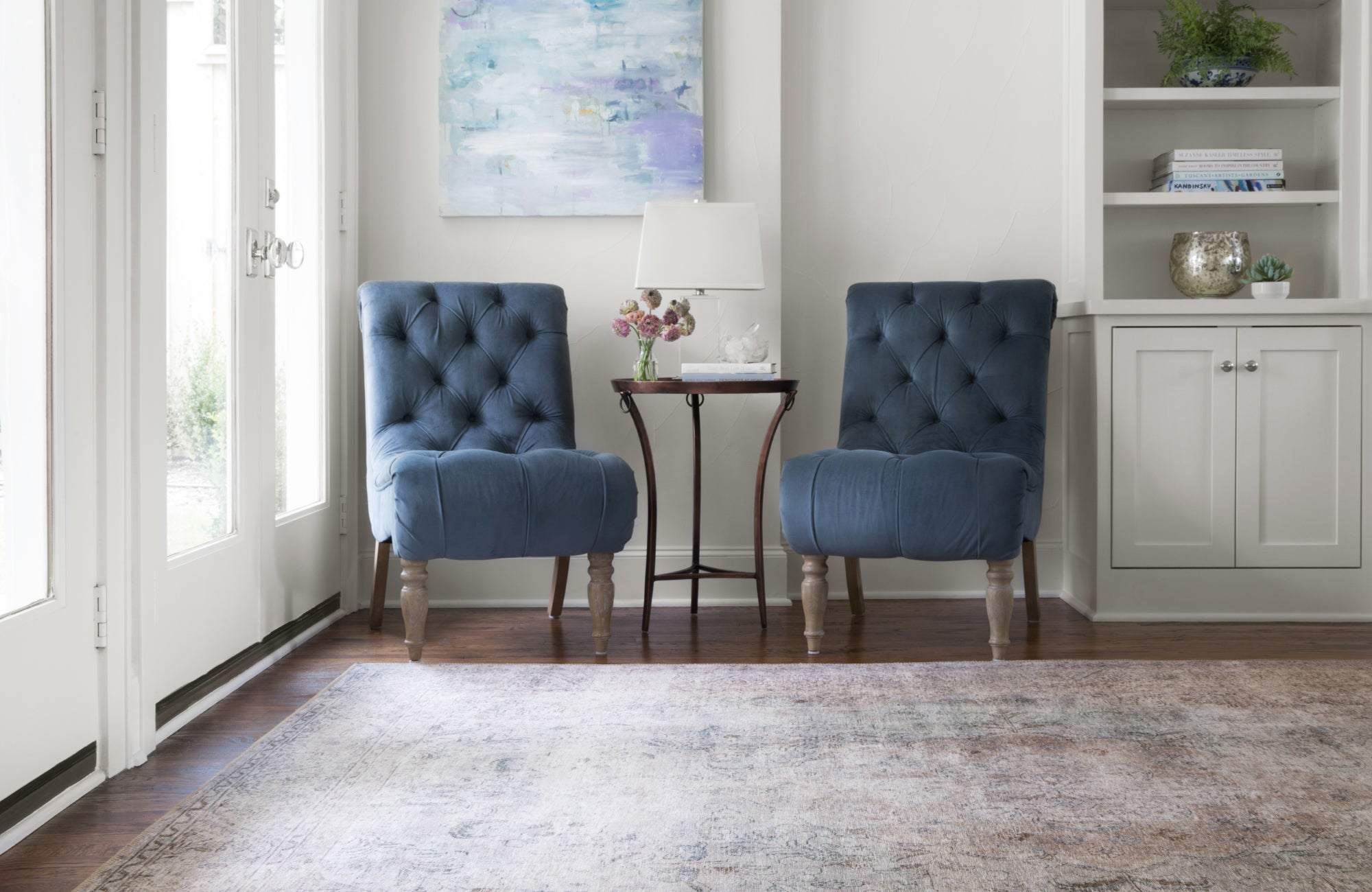 Living room corner with soft neutral tones featuring a 10x10 rug that grounds two blue tufted chairs beside tall windows.