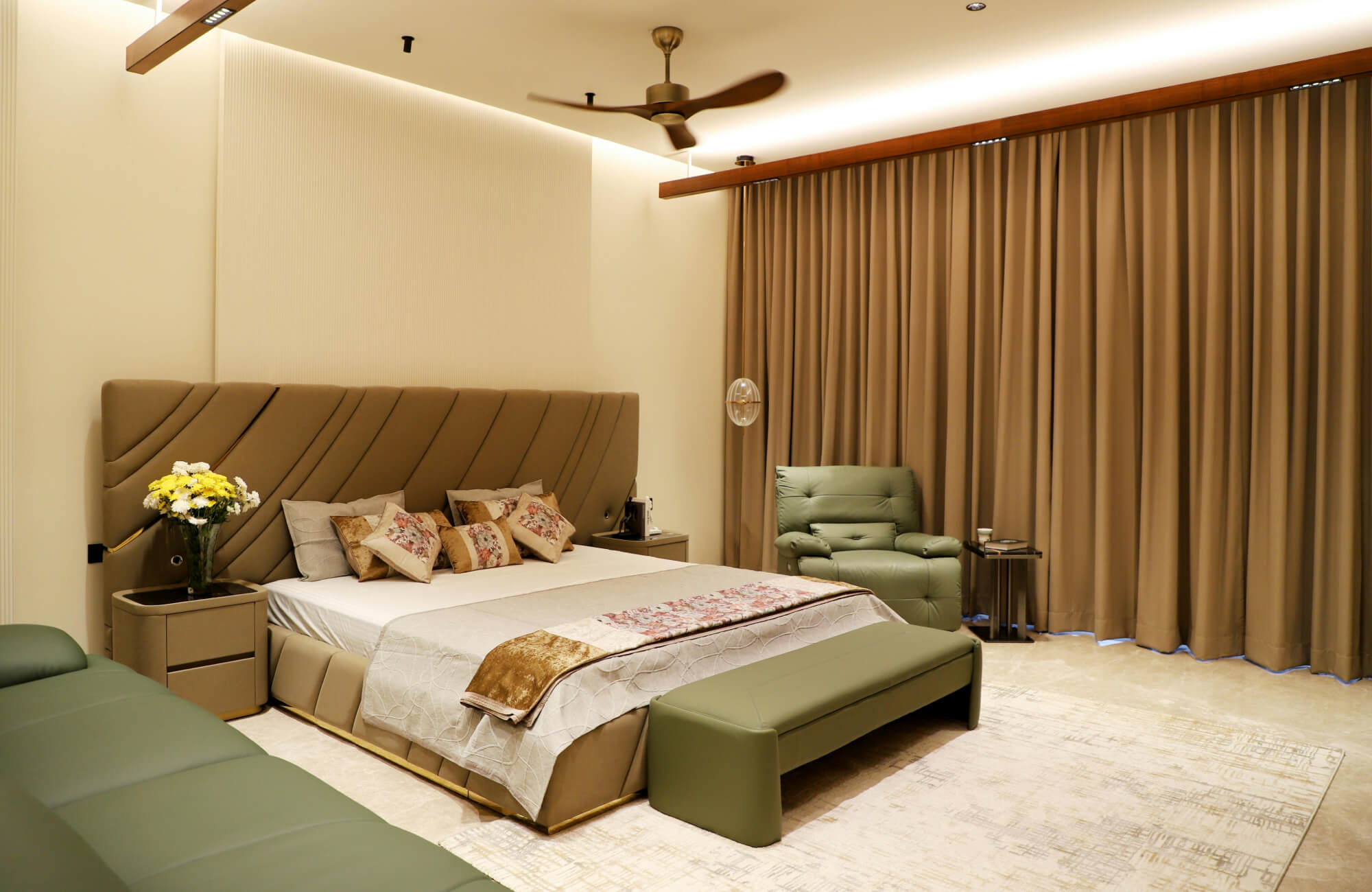 A warm, modern bedroom with an upholstered headboard, layered bedding, a green recliner and bench, and tall draped curtains adding softness to the room.