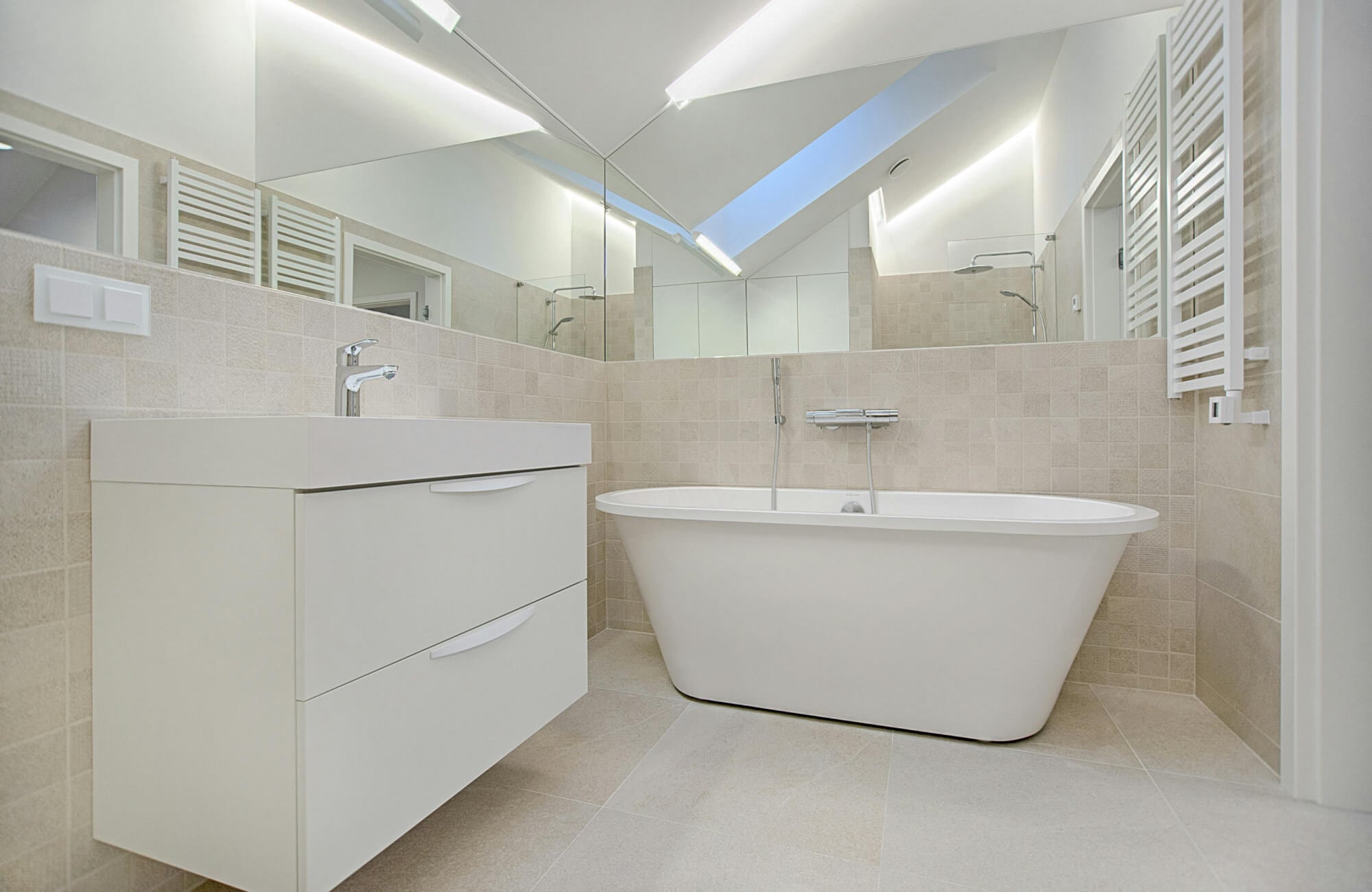 Bright, spa-like bathroom featuring neutral tile walls, sleek vanity drawers, and a contemporary soaking tub beneath a skylight.