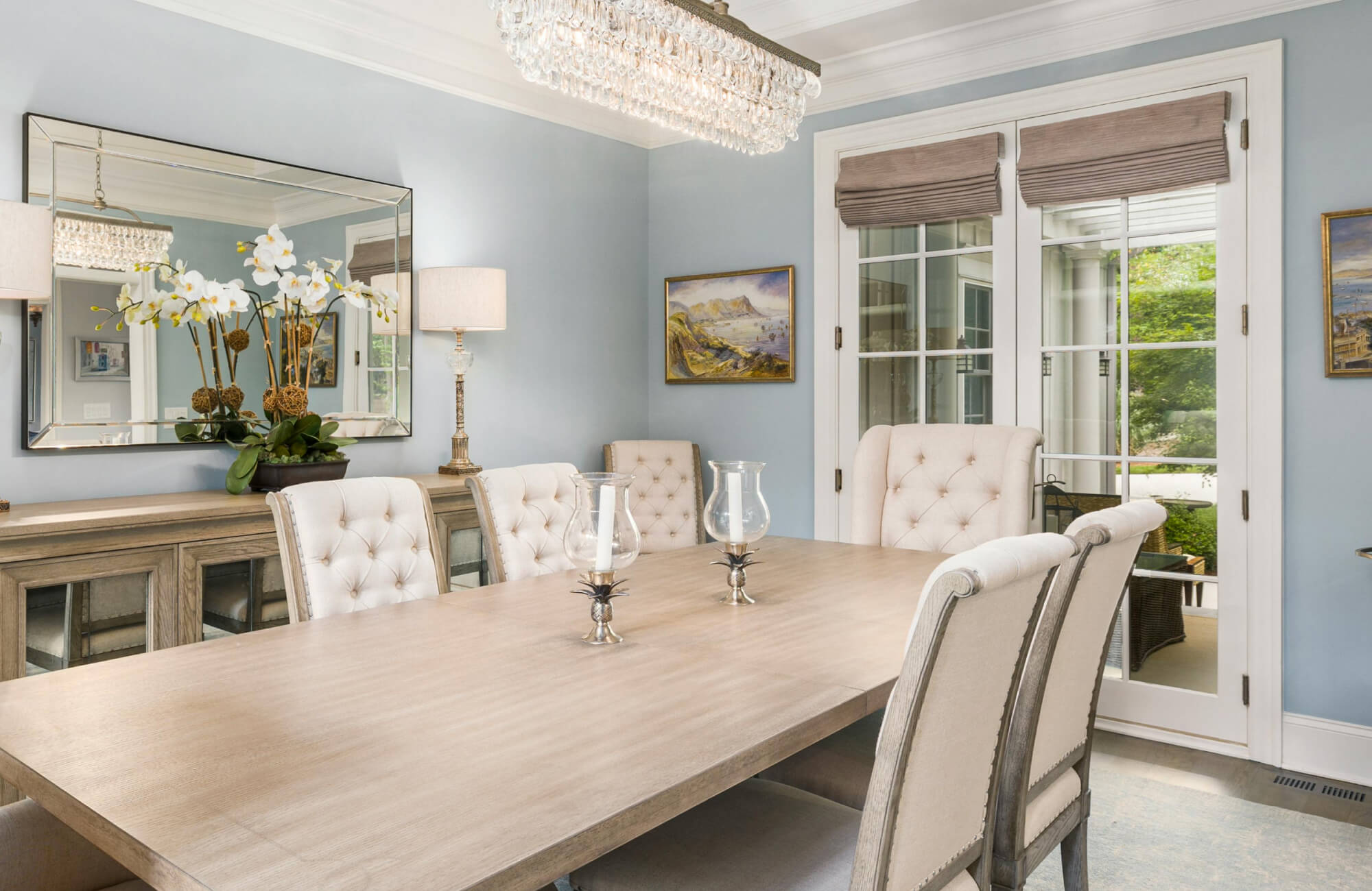 Refined dining space featuring a large rectangular dining room mirror above a wooden sideboard, reflecting a crystal chandelier, soft blue walls, tufted upholstered chairs, and elegant floral accents.
