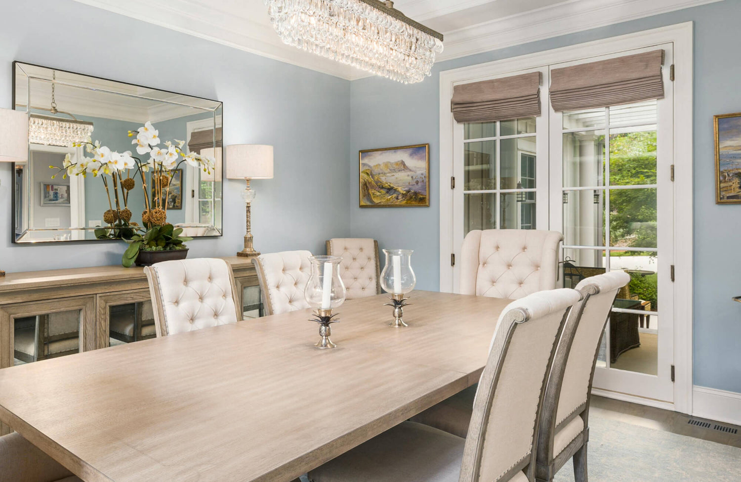 Refined dining space featuring a large rectangular dining room mirror above a wooden sideboard, reflecting a crystal chandelier, soft blue walls, tufted upholstered chairs, and elegant floral accents.