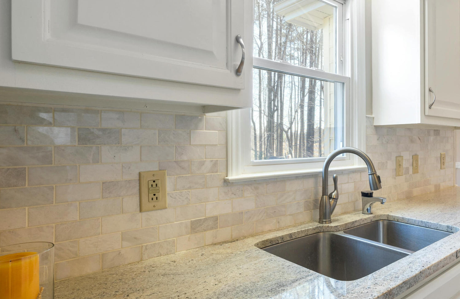Bright kitchen with a soft marble subway tile backsplash, granite countertops, and a stainless steel sink under a sunny window.