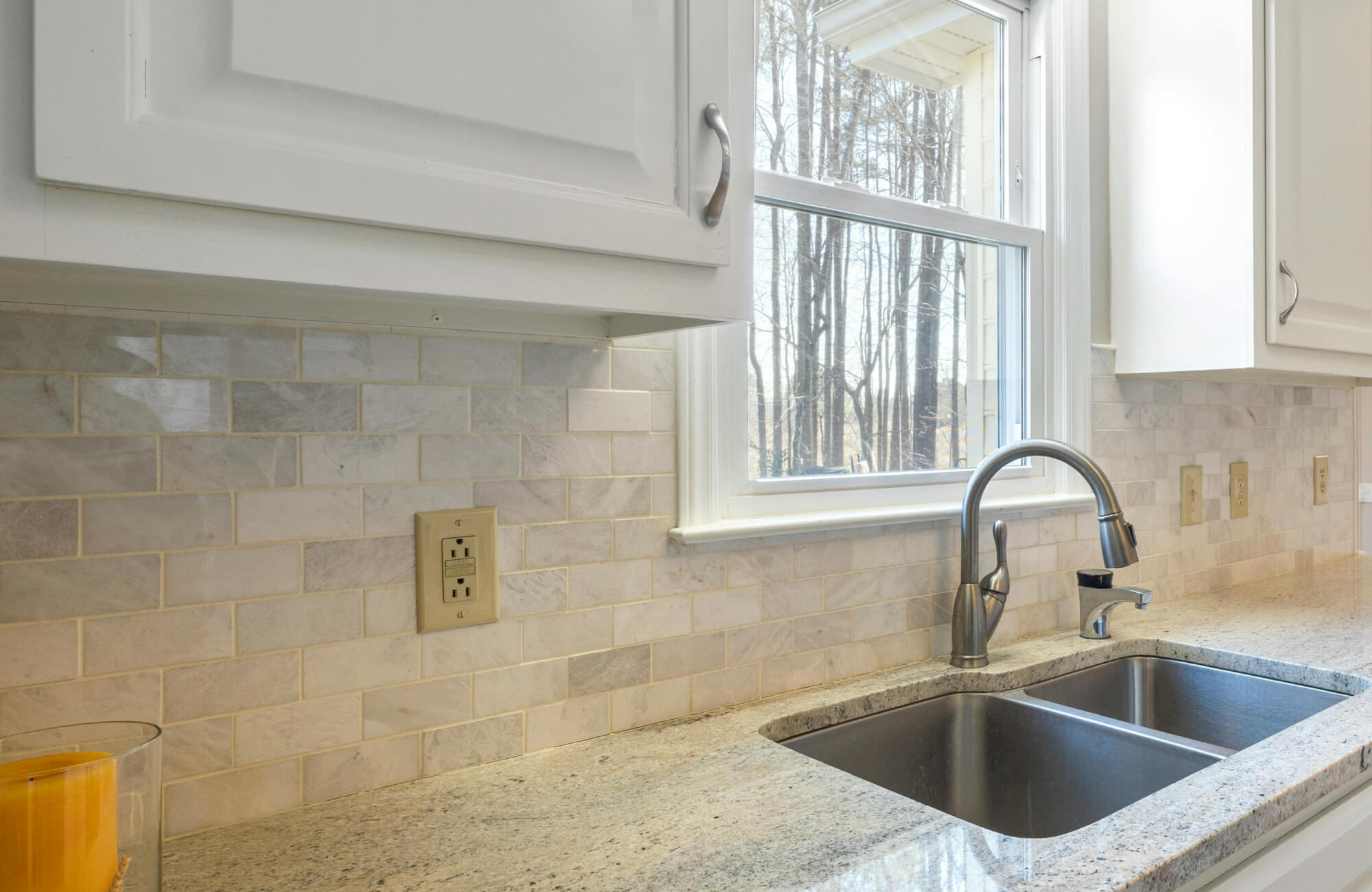 Bright kitchen with a soft marble subway tile backsplash, granite countertops, and a stainless steel sink under a sunny window.