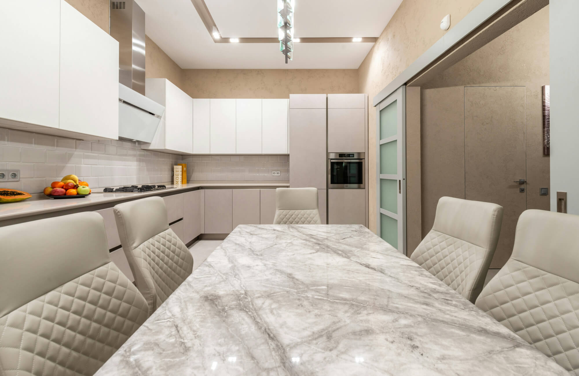 A polished gray marble dining table in a bright modern kitchen with white cabinetry and quilted dining chairs.