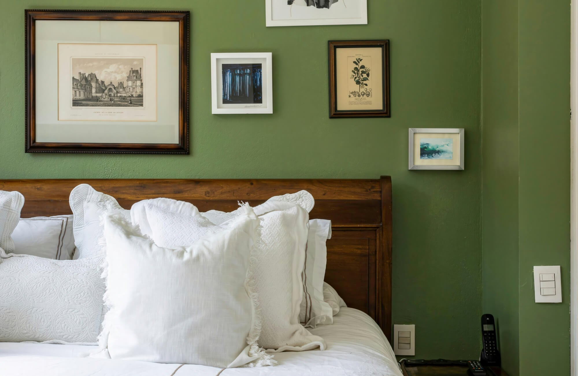 Gallery wall with mixed framed artwork above a wooden bed against a deep green wall, creating a layered focal point in a cozy bedroom.