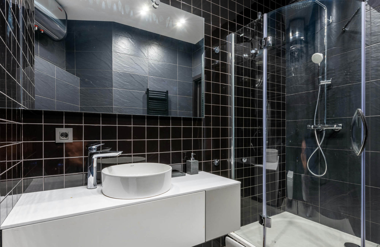Modern bathroom with black tiled shower and white vanity featuring reflective surfaces and bright overhead lighting.