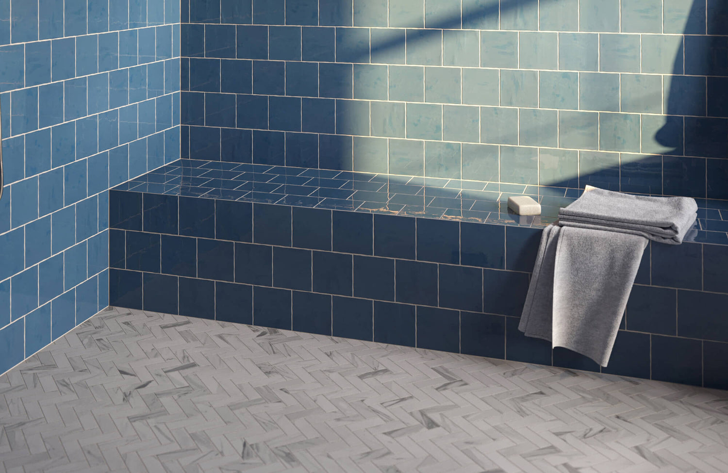 Serene bathroom shower featuring glossy blue wall tiles with a built-in bench, paired with a light gray herringbone tile floor that adds subtle pattern, texture, and modern elegance to the space.