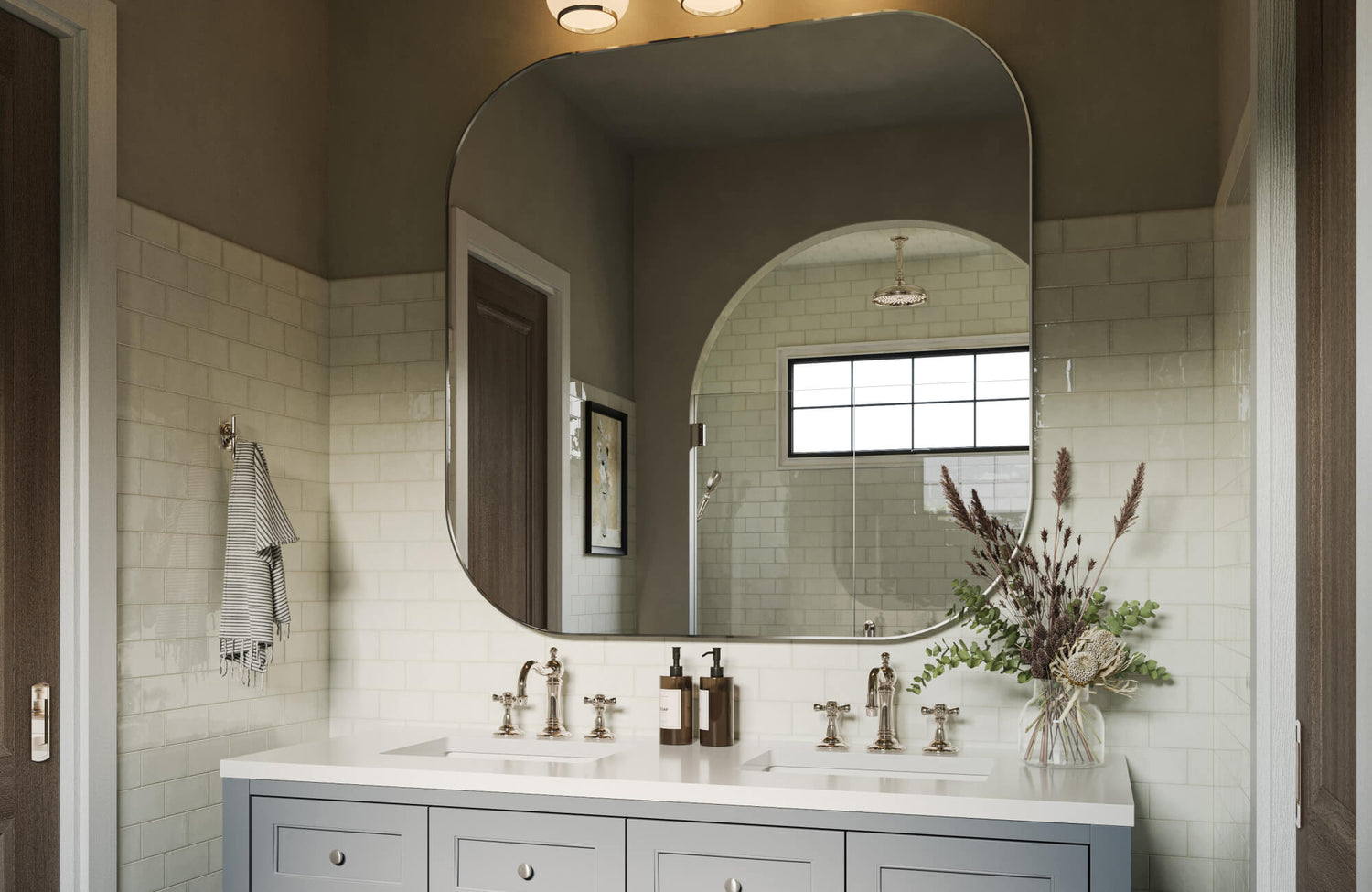 A bright, elegant bathroom features a soft gray double vanity topped with polished fixtures, glossy subway-tiled walls, and a striking large wall mirror that reflects the arched shower entry and enhances the room’s airy, sophisticated ambiance.
