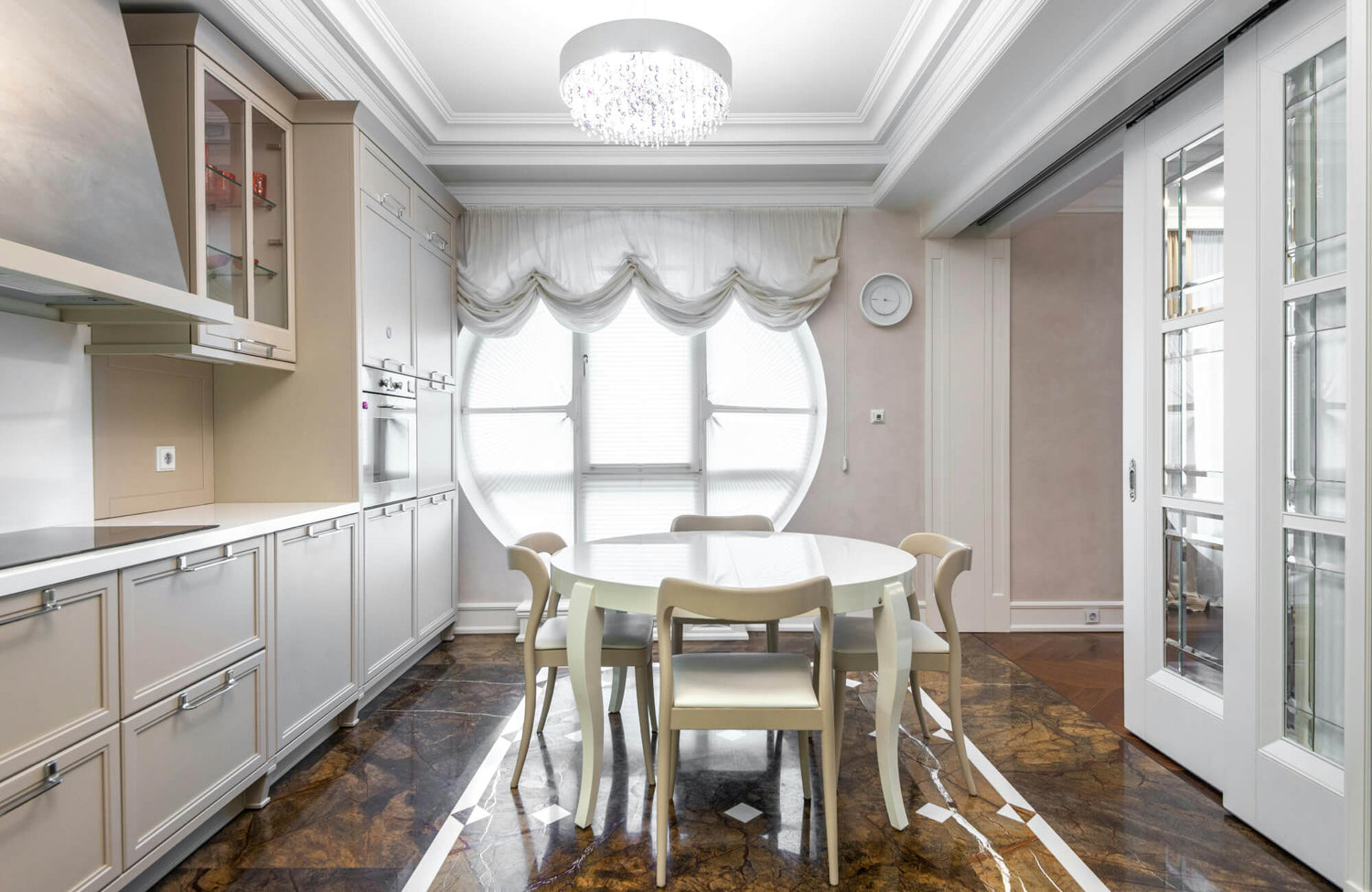 Alt text: Elegant kitchen dining area with glossy brown marble flooring, white cabinetry, a round white table with four chairs, and a large circular window dressed in soft drapery.