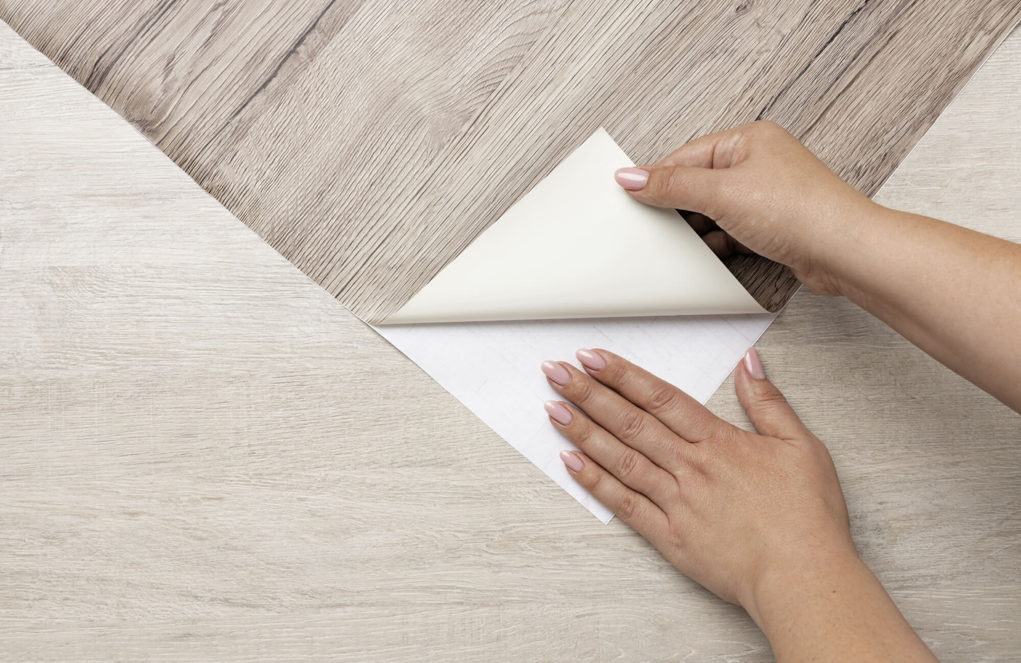 Hands applying peel-and-stick flooring with a light wood grain design over a smooth surface, showing the adhesive backing being pressed down.