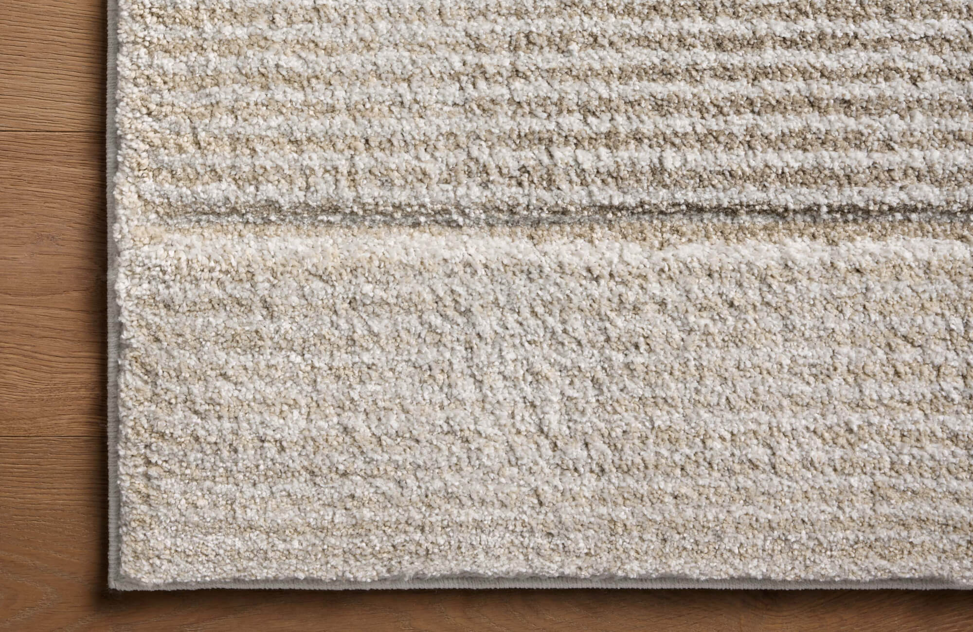 Close-up of a plush polypropylene rug with a soft, striped texture in neutral beige tones, laid over warm wood flooring.