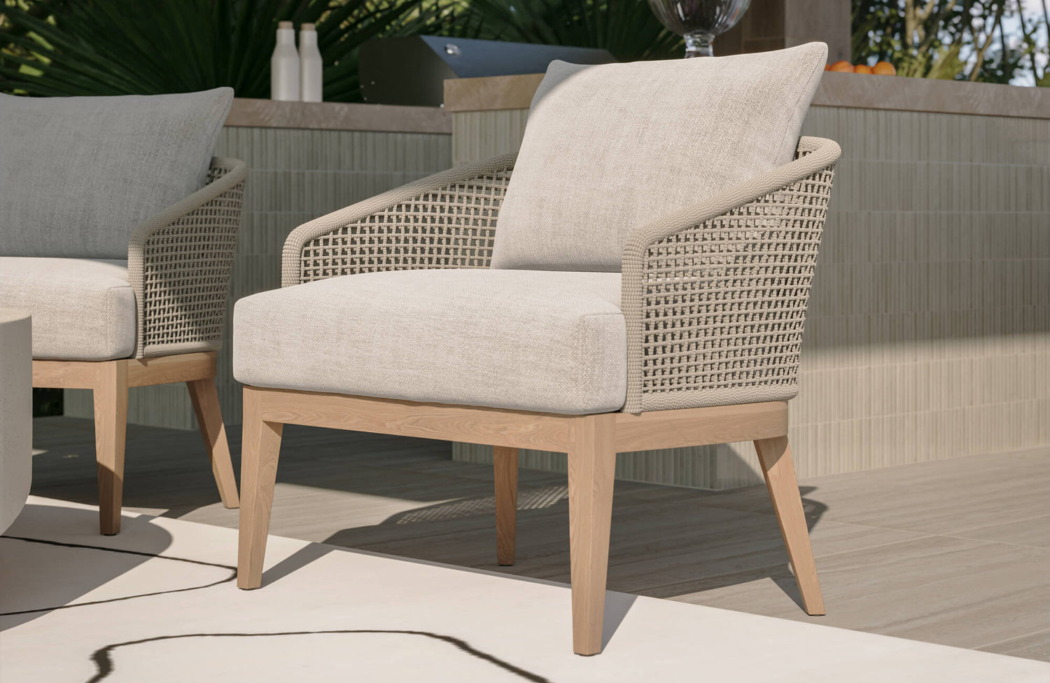 Outdoor lounge chair with woven rope arms, cream cushions, and a light wood frame beside a tiled patio bar.