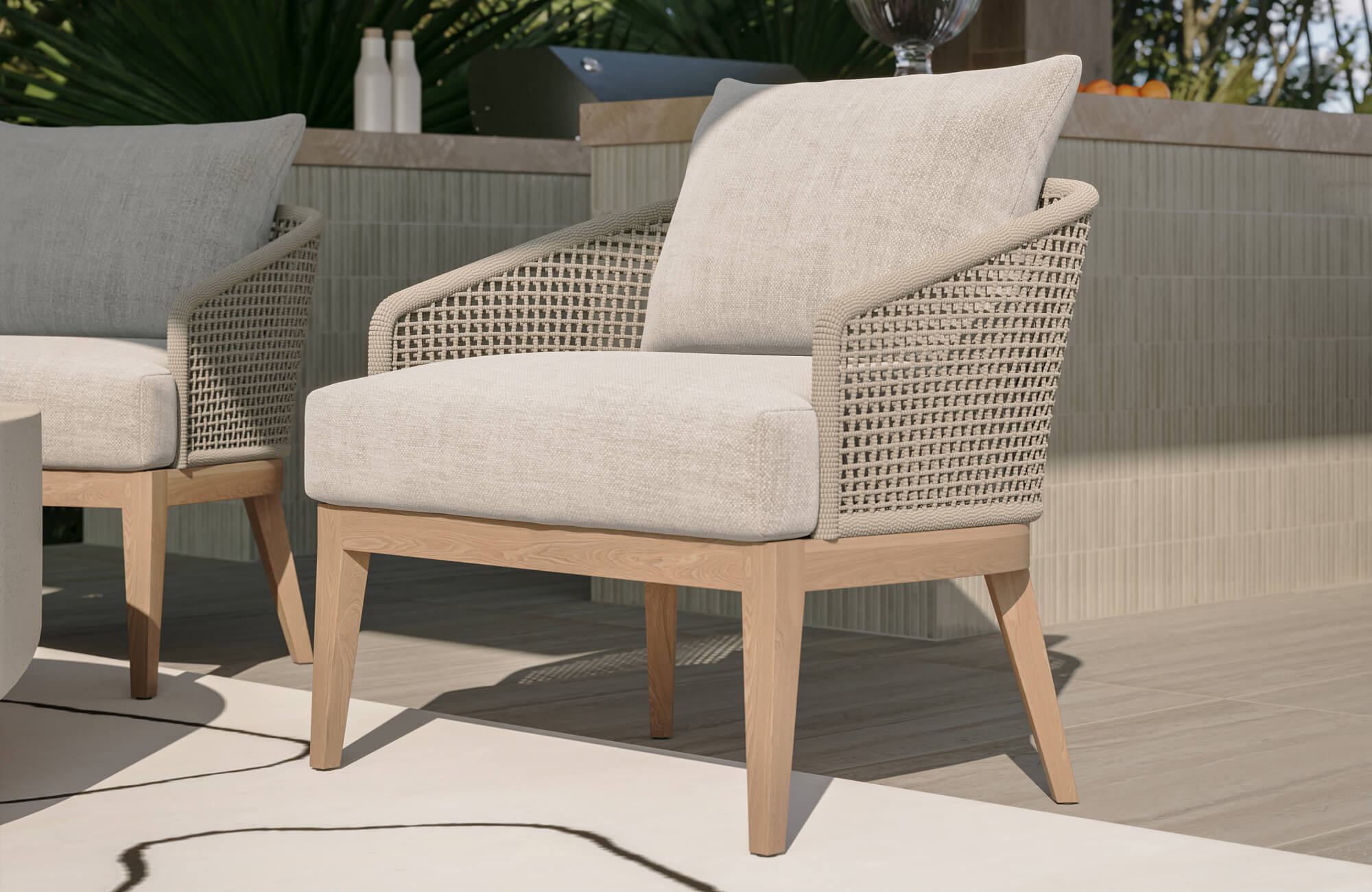 Outdoor lounge chair with woven rope arms, cream cushions, and a light wood frame beside a tiled patio bar.