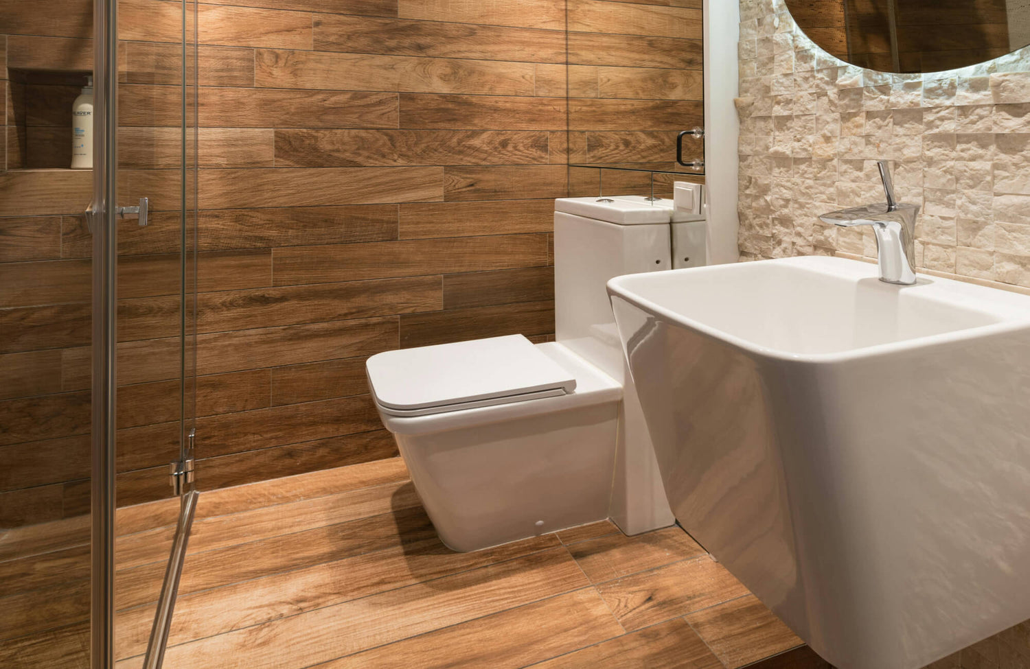 A contemporary bathroom features rich wood looking tile on both the floor and walls, creating a warm, natural backdrop that contrasts beautifully with sleek white fixtures and textured stone accents.