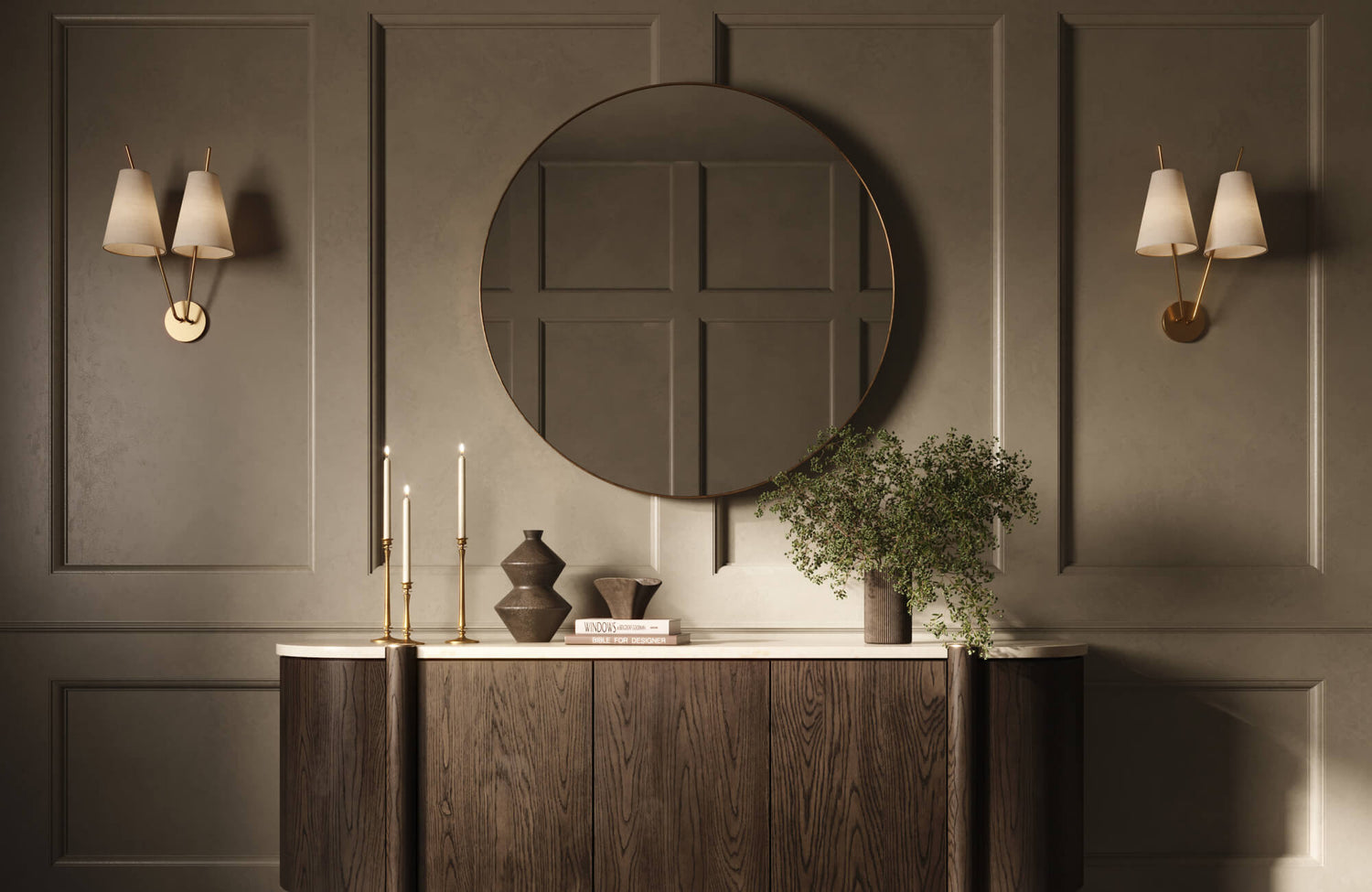A large round mirror hangs above a dark wood console styled with candles, books, and greenery, set against paneled taupe walls with warm brass wall sconces.