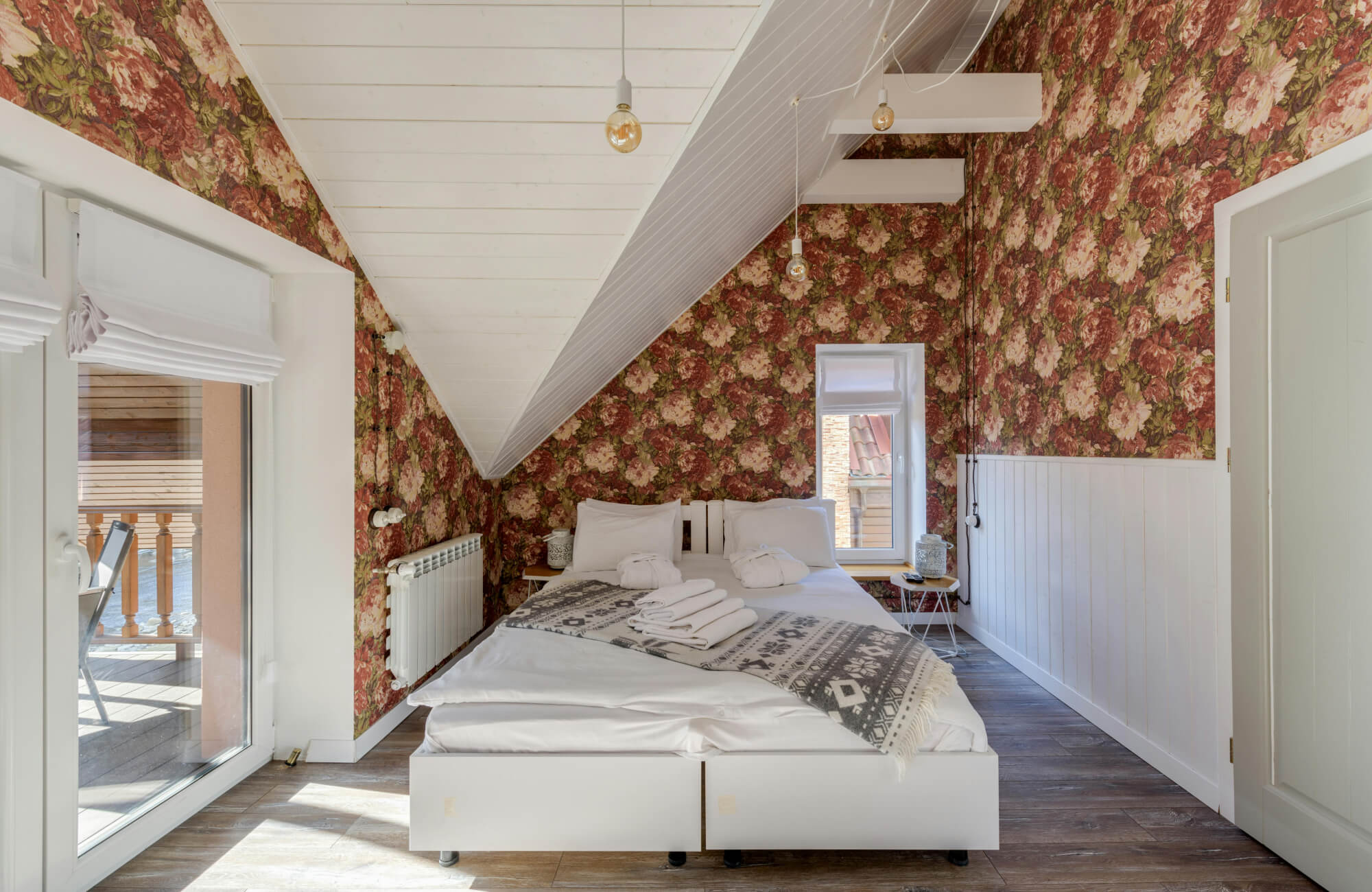 Cozy attic bedroom with red floral wallpaper, white sloped ceiling, and warm wood look tile flooring under a simple bed.