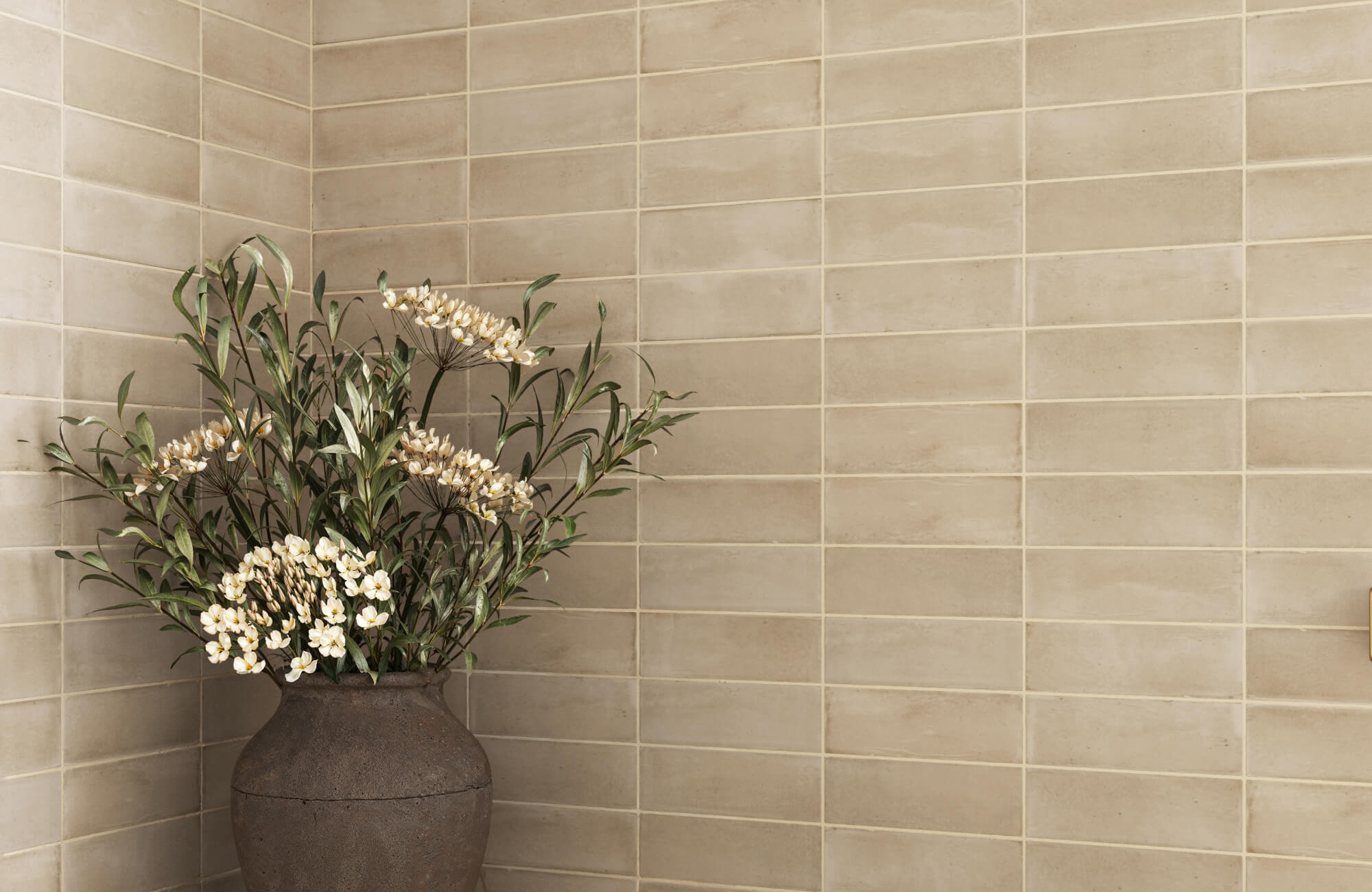 Matte subway tiles in a warm neutral tone line the corner wall, accented by a rustic vase with white flowers and greenery.