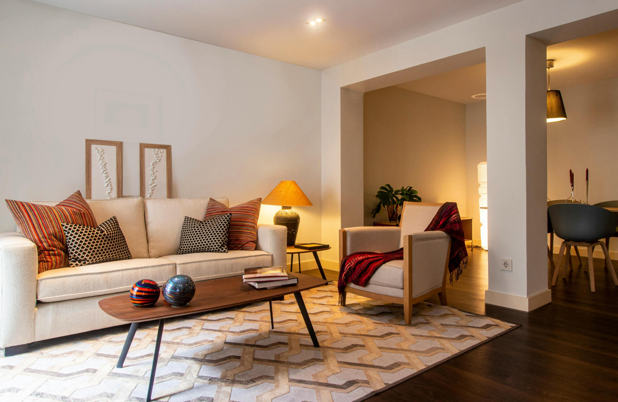 Cream-colored sofa and armchair set atop a geometric beige and taupe rug, accented with patterned cushions and warm lighting.