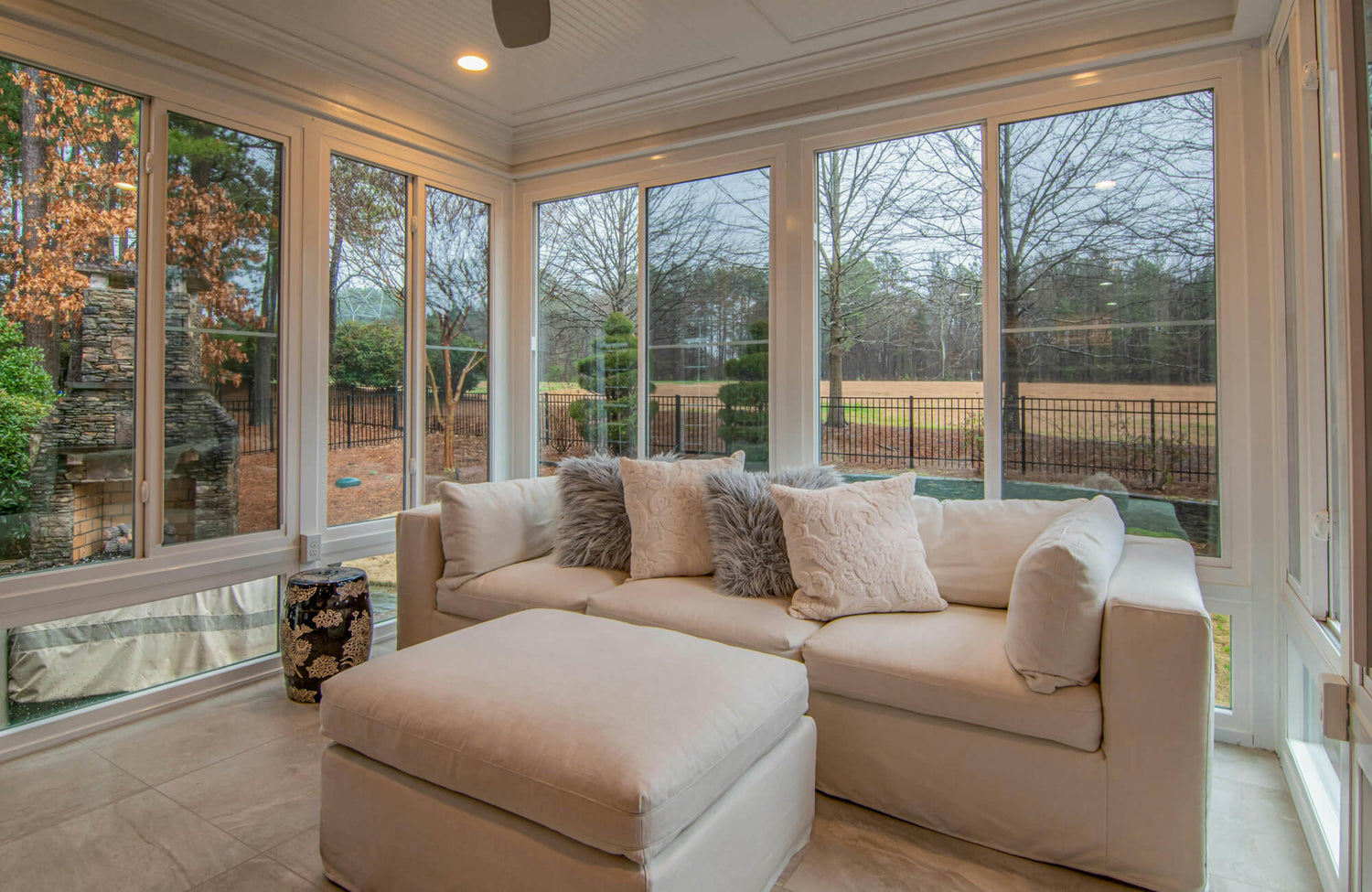 Bright sunroom with large picture windows, beige floor tiles, and plush sofa styled with textured and faux fur decorative pillows.