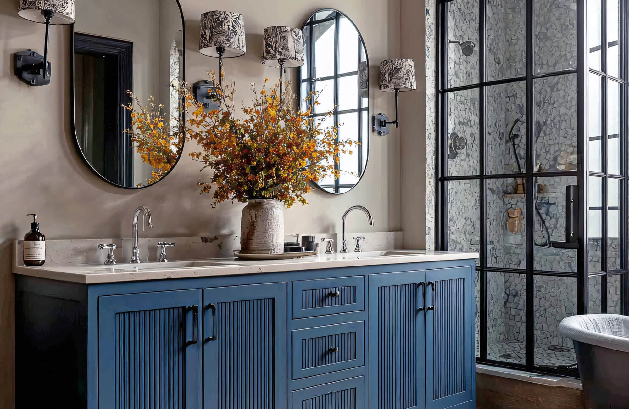 Blue double-sink vanity with two oval mirrors, patterned sconces, marble shower, and floral arrangement in an elegant bathroom.
