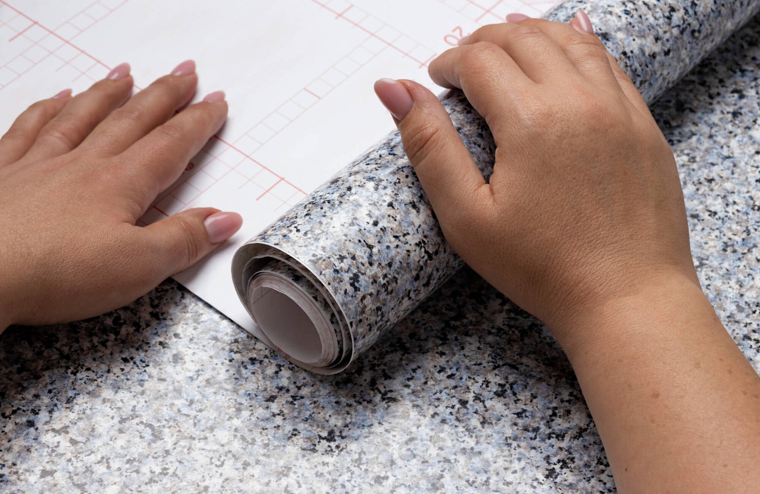 Hands unrolling granite-look wallpaper with grid backing, applying peel-and-stick wallpaper over a countertop surface for a smooth, seamless finish.