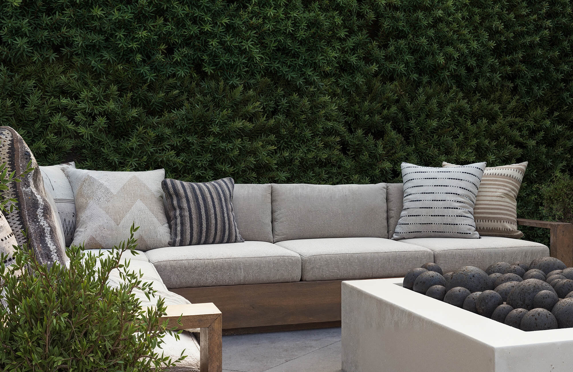 Decorative pillow arrangement on a neutral outdoor sectional sofa with textured patterns and a greenery backdrop.