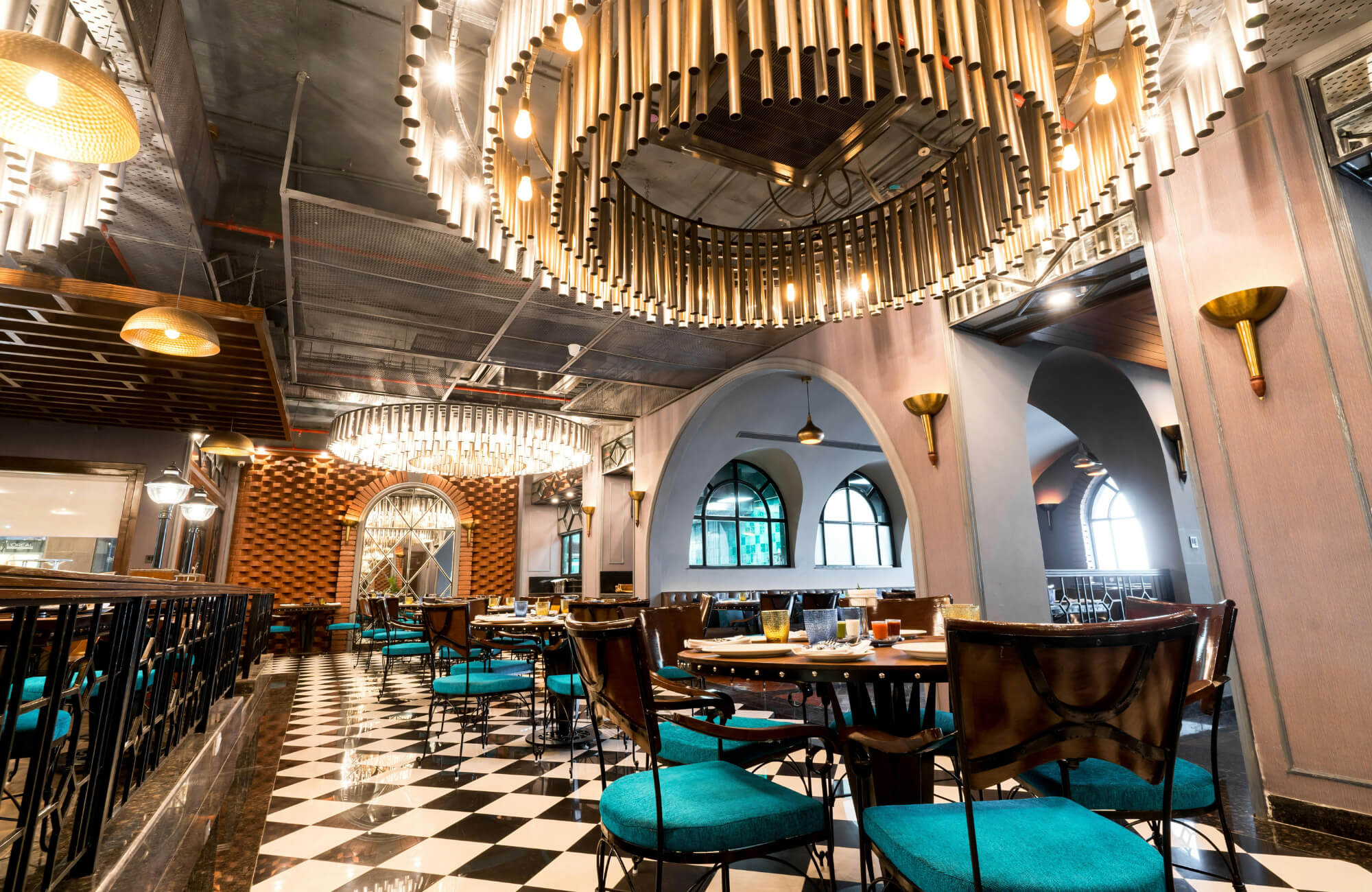 Elegant restaurant interior featuring black-and-white chequered tile flooring, modern lighting, arched windows, and teal-upholstered chairs.