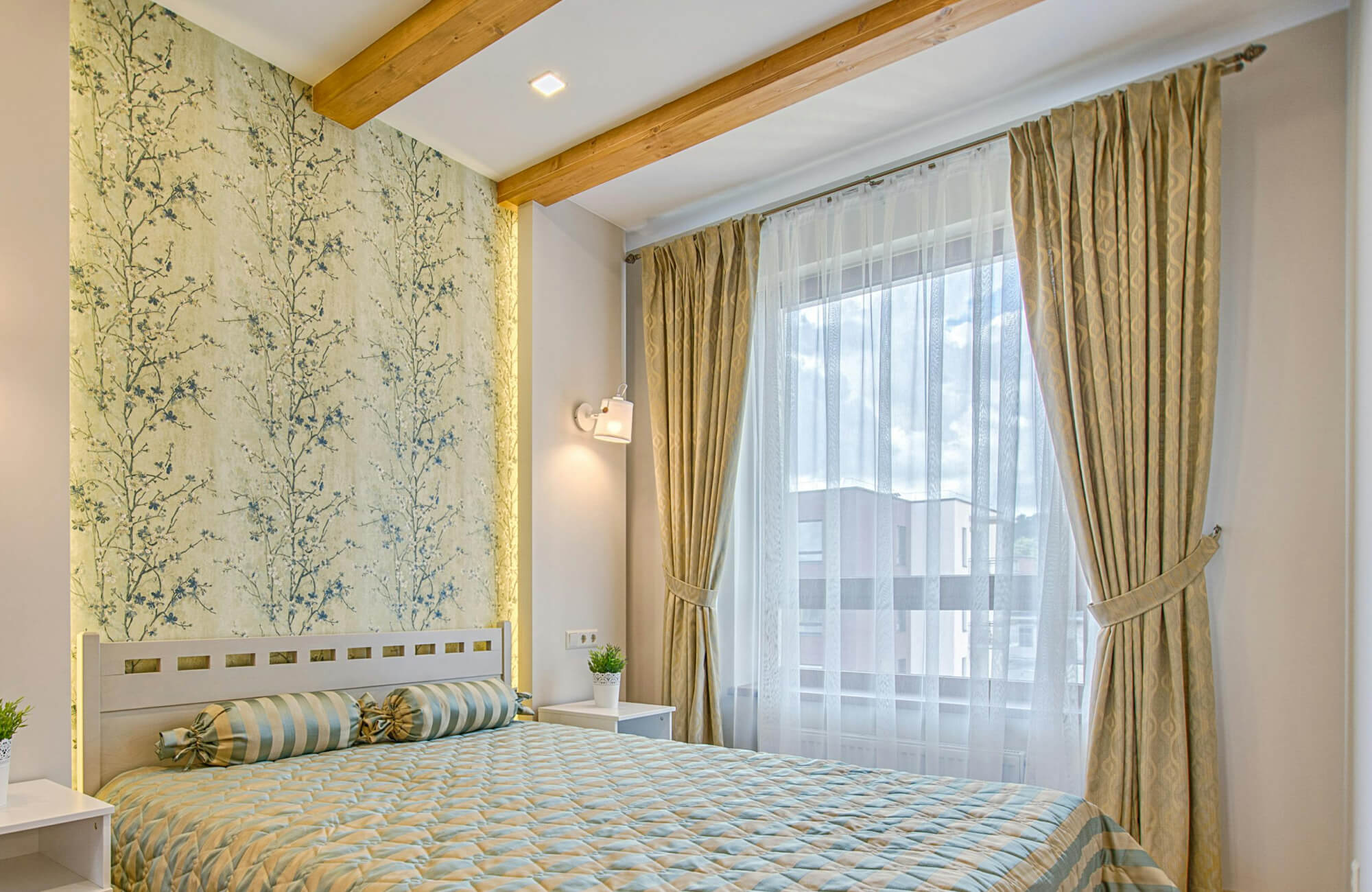 Bedroom featuring botanical wallpaper with delicate blue floral branches on a soft neutral background, behind the upholstered bed, and complements the warm wood ceiling beams and natural light from the window.