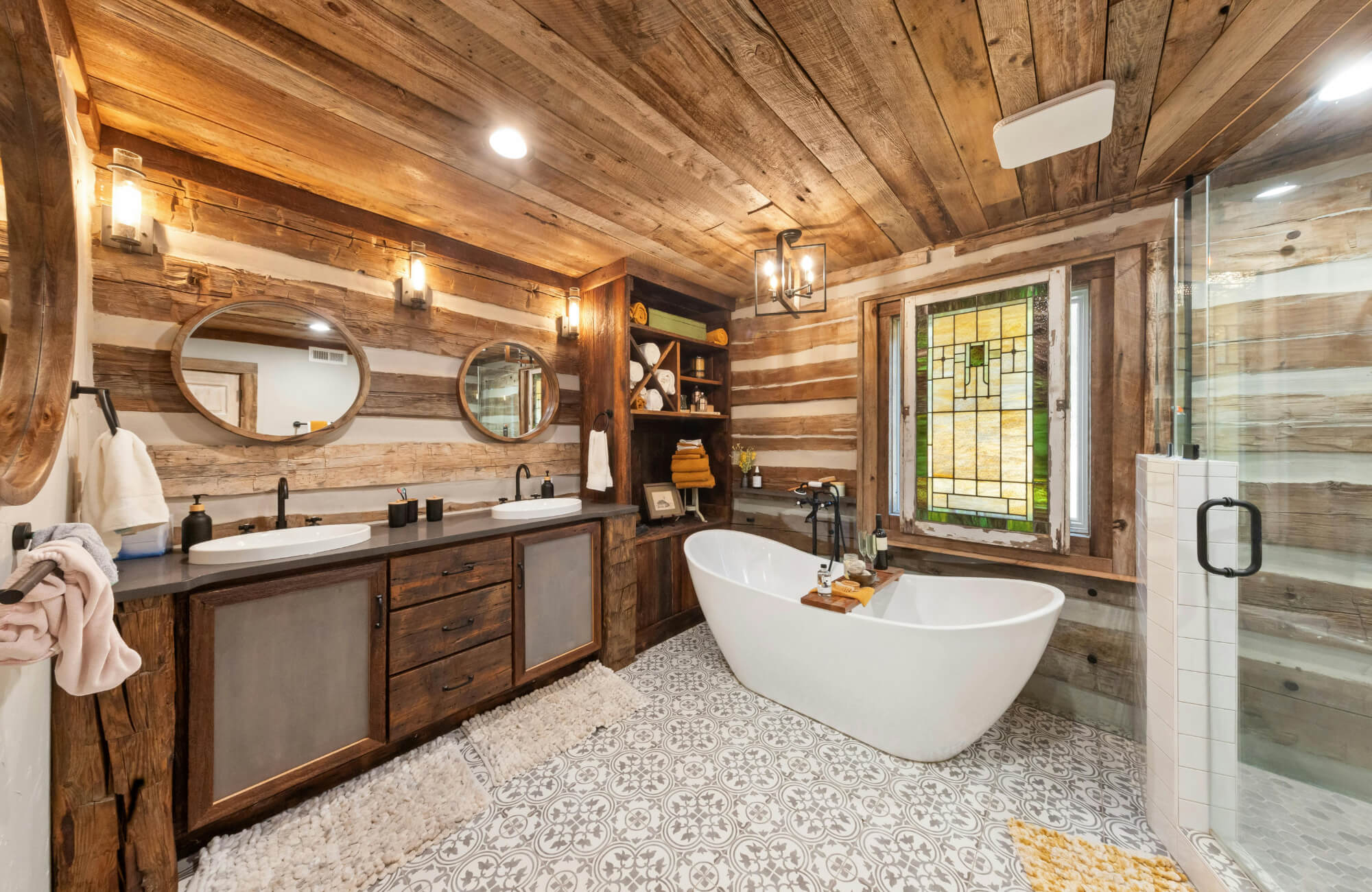 Rustic bathroom with double-sink vanity, wood panel walls and ceiling, freestanding tub, patterned tile floor, and stained-glass window.