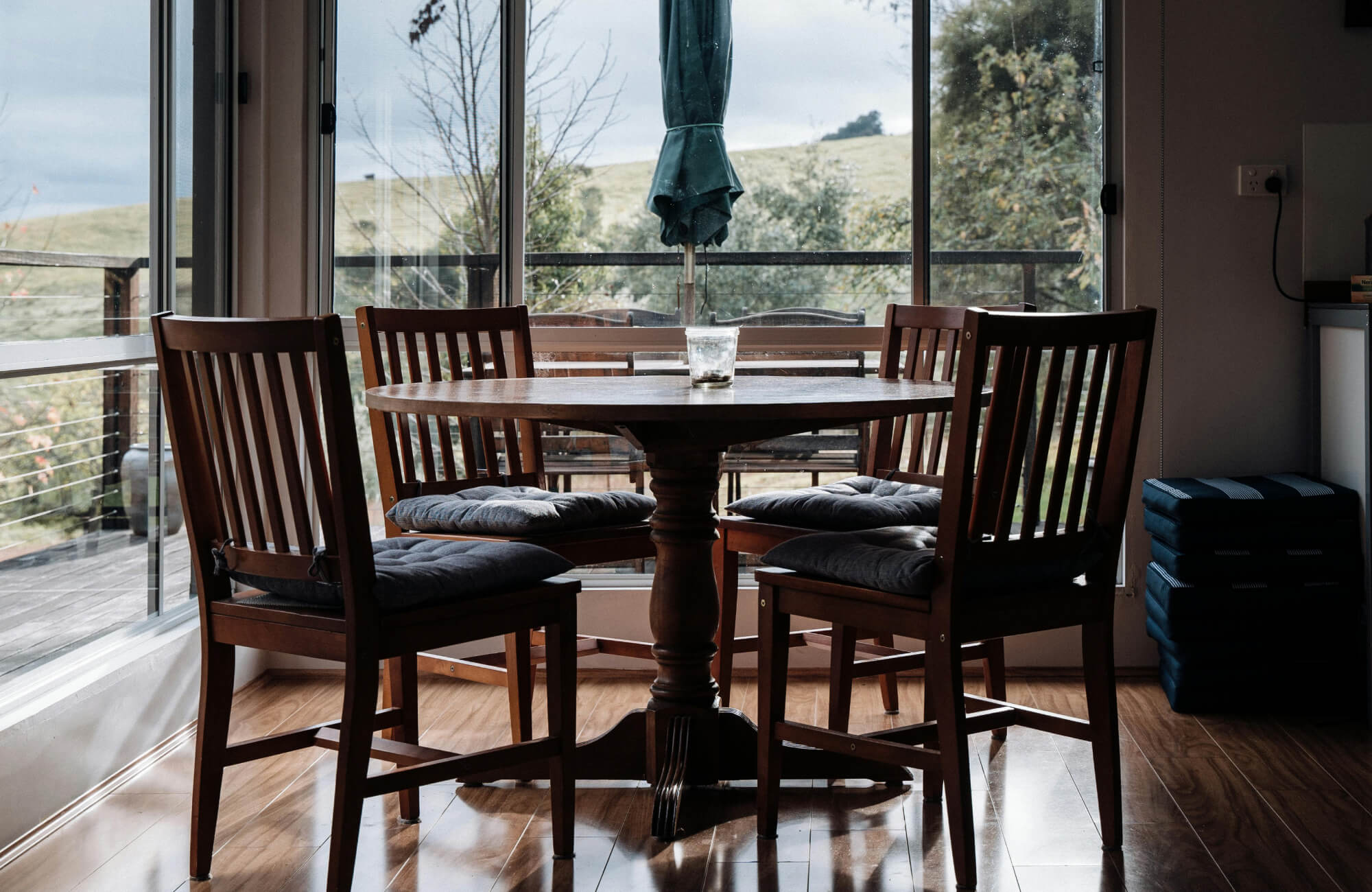 A warm, sunlit dining nook features a wooden round dining table set with four cushioned chairs, framed by large glass doors that open to a serene outdoor view.