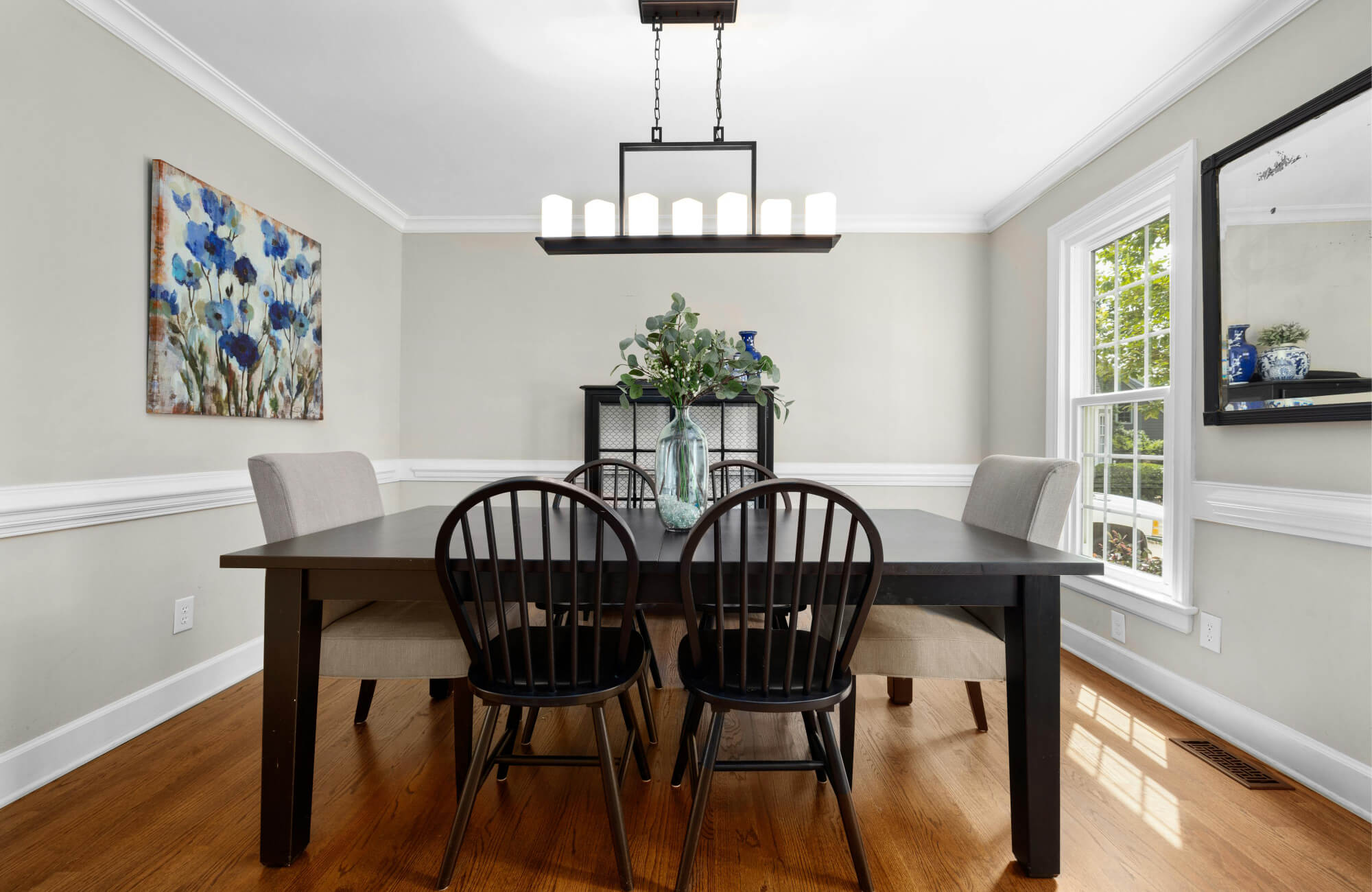 Black dining table centered on warm wood floors, paired with mixed seating and natural light from large windows to keep the room open and balanced.