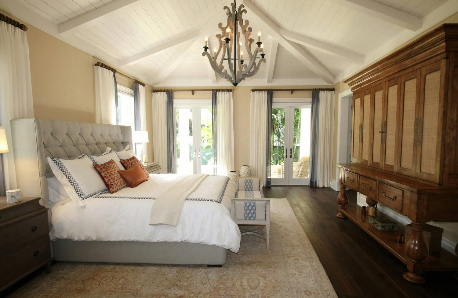 Airy bedroom with dark wood look tile flooring, a cream patterned rug, a tufted headboard, and a vaulted white ceiling with French doors.