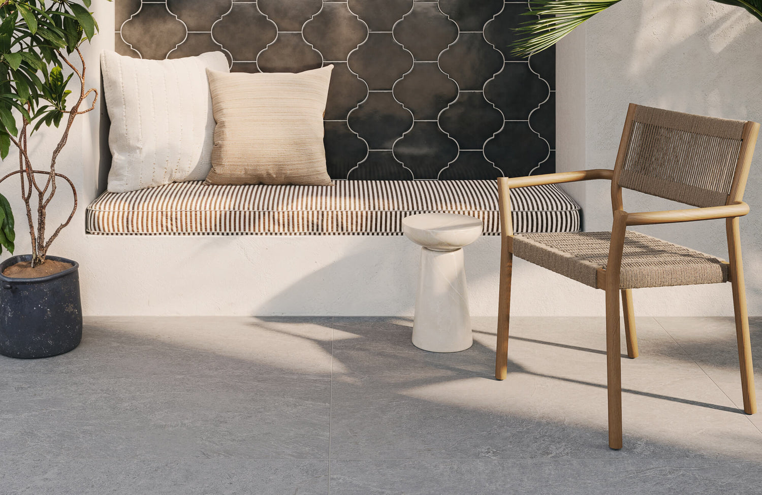 Outdoor seating nook featuring light gray paver tiles with a smooth stone texture, paired with a black arabesque tile backsplash, a striped cushioned bench, and natural wood furniture that evokes a serene, modern Mediterranean style.