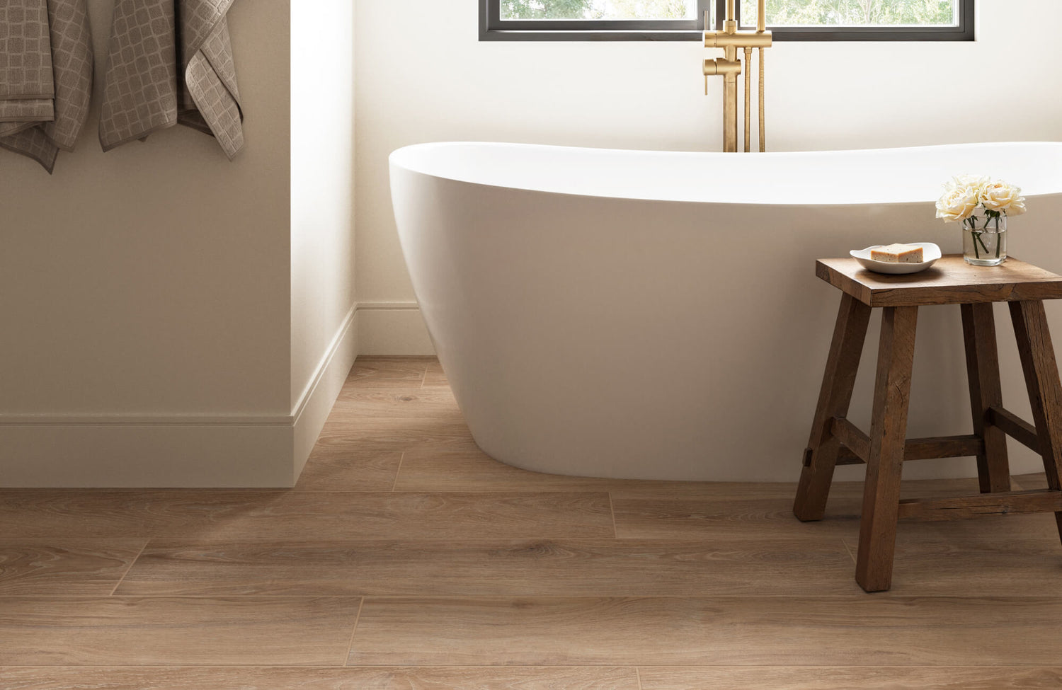 A serene bathroom featuring warm wood-look porcelain tile flooring, a sleek white freestanding tub, and elegant brass fixtures.
