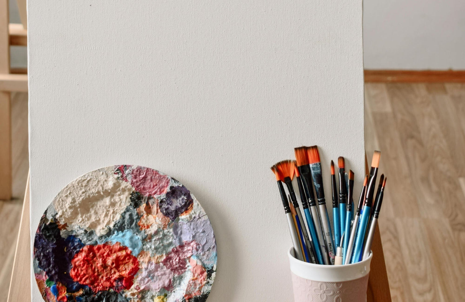 Canvas painting wall art setup featuring a blank canvas on an easel with a textured paint palette and assorted brushes in a bright studio setting.