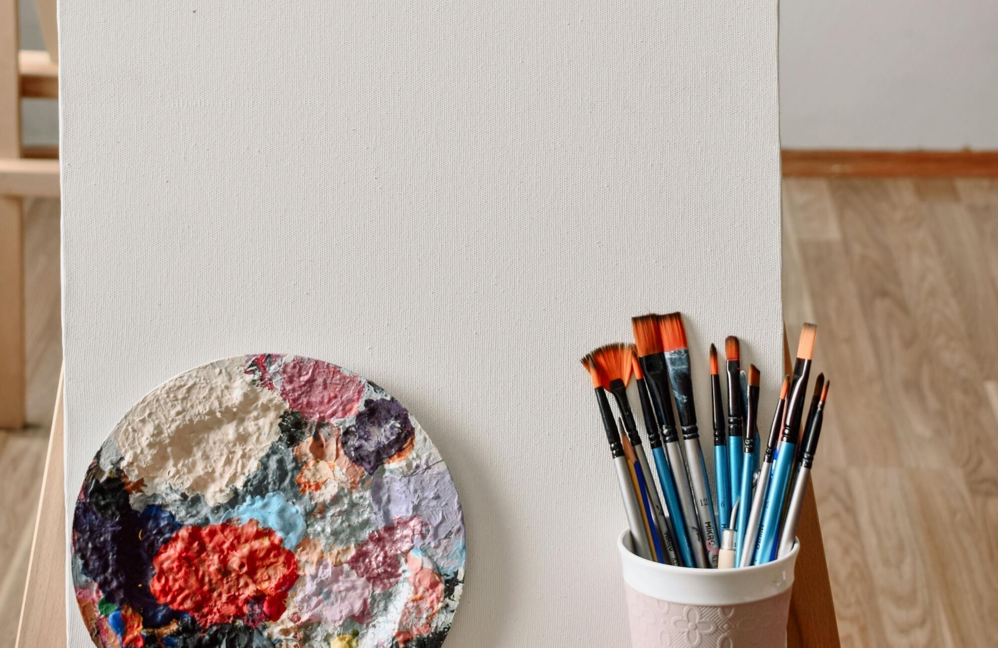 Canvas painting wall art setup featuring a blank canvas on an easel with a textured paint palette and assorted brushes in a bright studio setting.