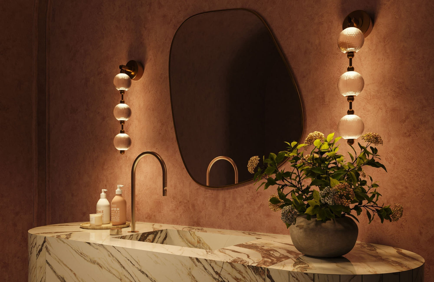 Moody bathroom setting featuring an asymmetrical mirror, textured wall finish, brass globe sconces, and a marble vanity styled with greenery and warm light.
