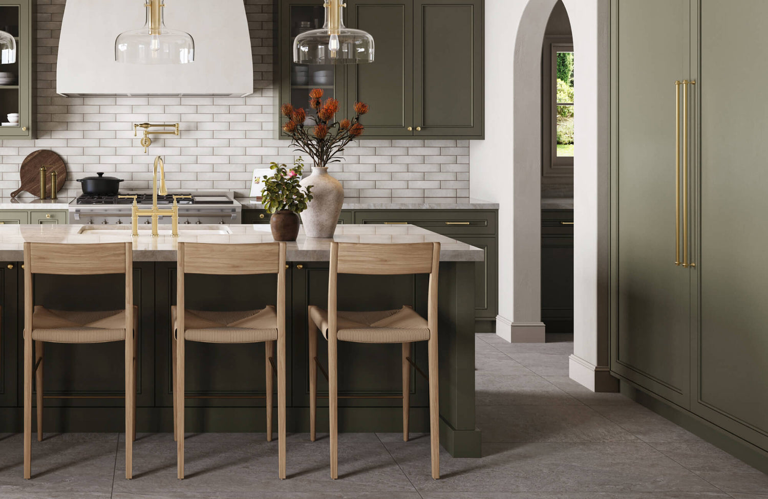 A warm, sophisticated kitchen features an olive-toned island paired with light wood modern counter stools, set against a backdrop of textured subway tile, brass accents, and elegant glass pendant lighting.