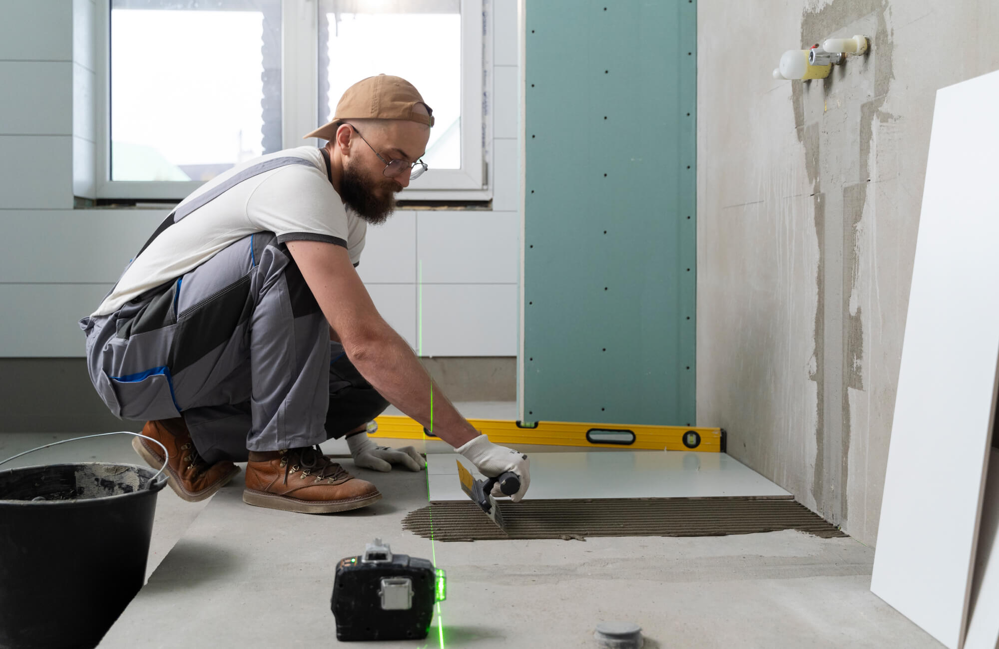 A contractor spreads tile adhesive on a bathroom floor, using a trowel and laser level to prep the surface for precise tile installation.