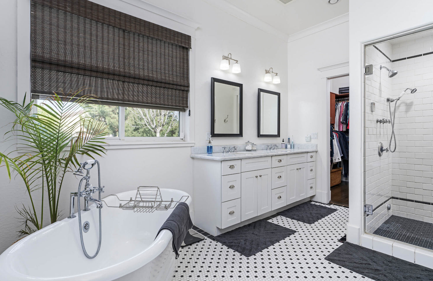 Bright bathroom with a freestanding tub, double vanity, walk-in shower, and black-and-white mosaic tile flooring with black mats.