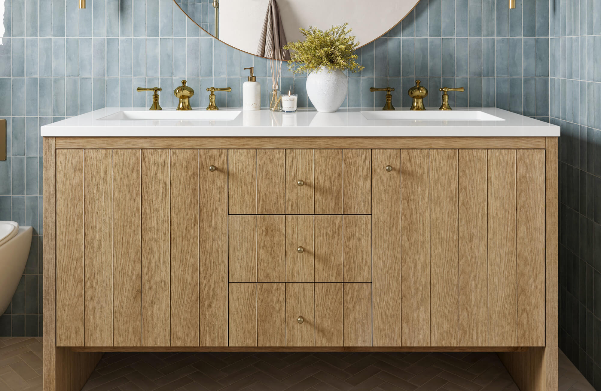 Light oak double vanity with white countertop and gold hardware, styled with matching brass faucets and a round mirror against soft blue tile walls.