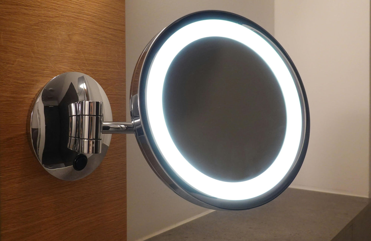 A close-up of a wall-mounted LED makeup mirror with a bright circular light, featuring a chrome arm and base against a warm wood panel.