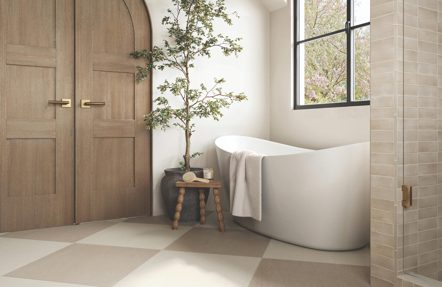 Serene bathroom with checkerboard floor tiles, freestanding tub, indoor tree, wooden doors, and neutral shower wall tiles.