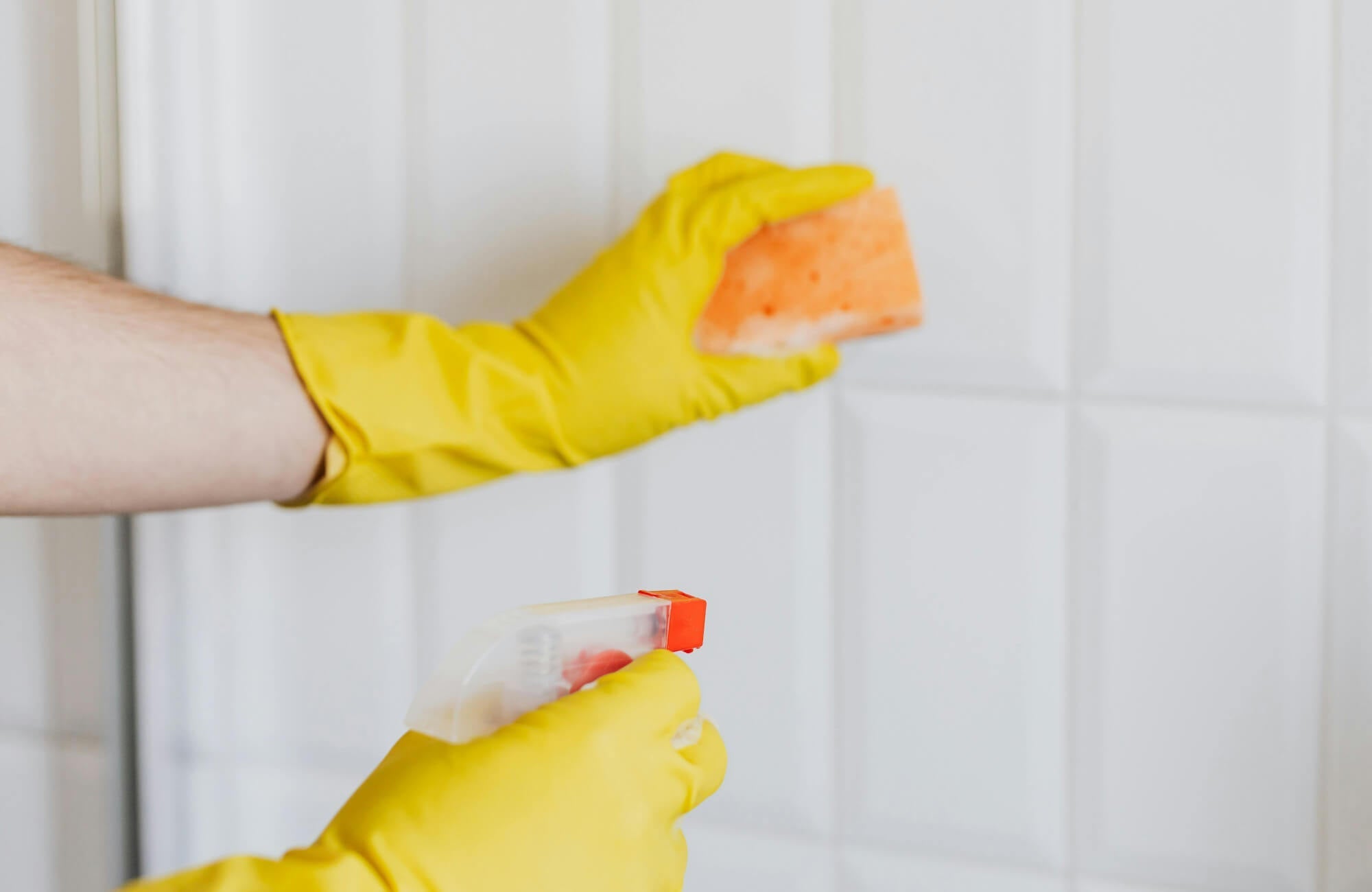 Yellow-gloved hands spraying cleaner and holding a sponge against white bathroom subway tiles, showing routine tile cleaning near grout lines.