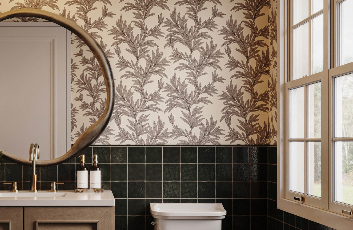 Wallpaper with brown botanical leaf pattern above dark green bathroom tiles, paired with a round mirror and wood vanity near a window.