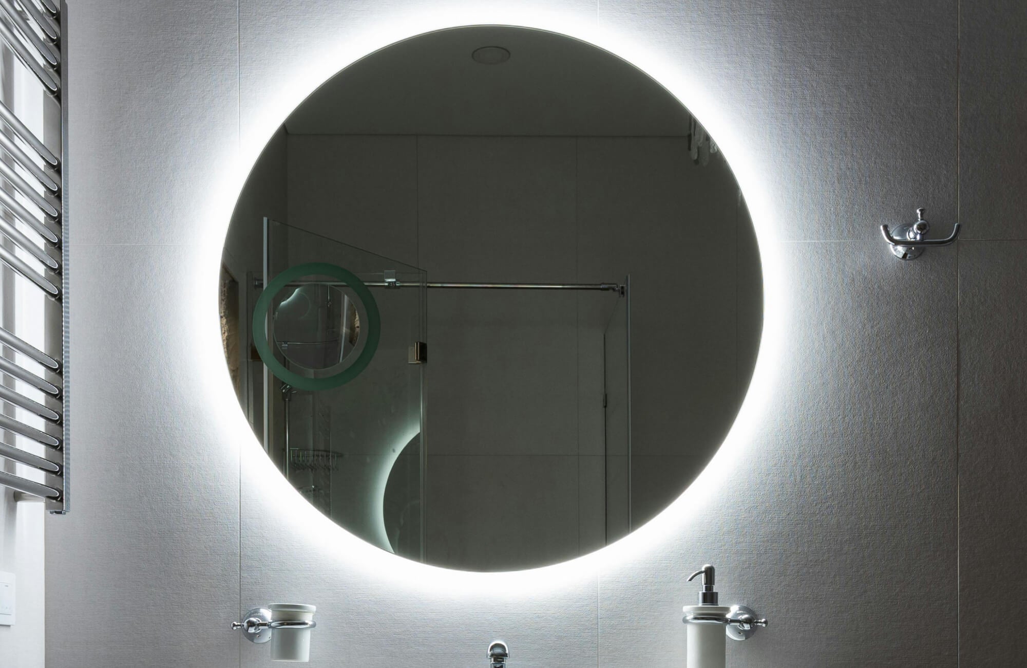 Contemporary bathroom wall featuring a glowing circle mirror with integrated LED backlighting, mounted against textured gray tiles and reflected by a glass shower enclosure and polished chrome accessories for a clean, modern look.