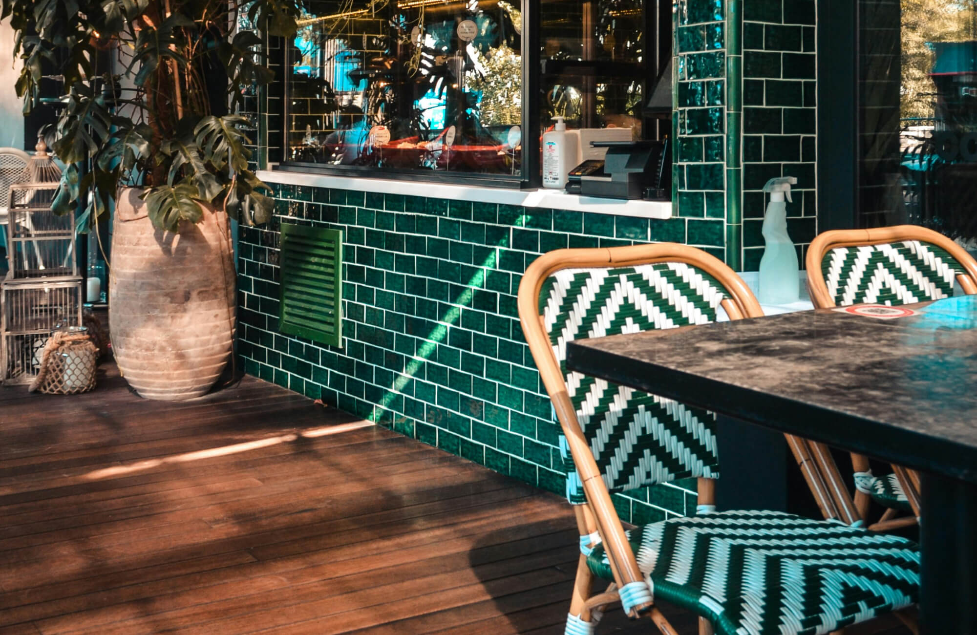 Glossy emerald green subway tiles contrast with woven chevron-patterned chairs, creating a vibrant and stylish café setting.