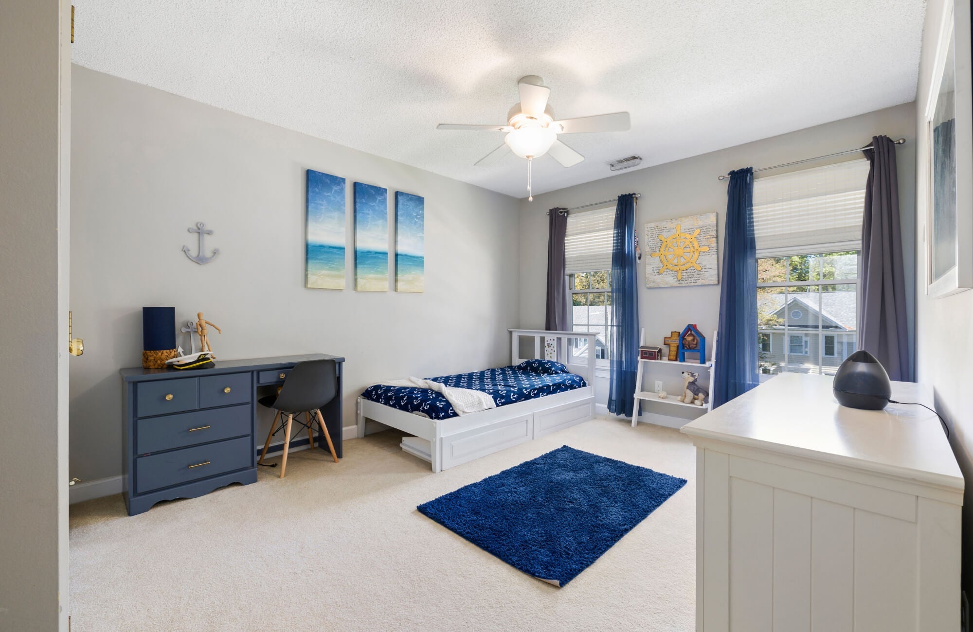 Nautical-themed bedroom with a navy blue rug, white bed, blue desk, and ocean artwork creating a bright, coastal-inspired space.