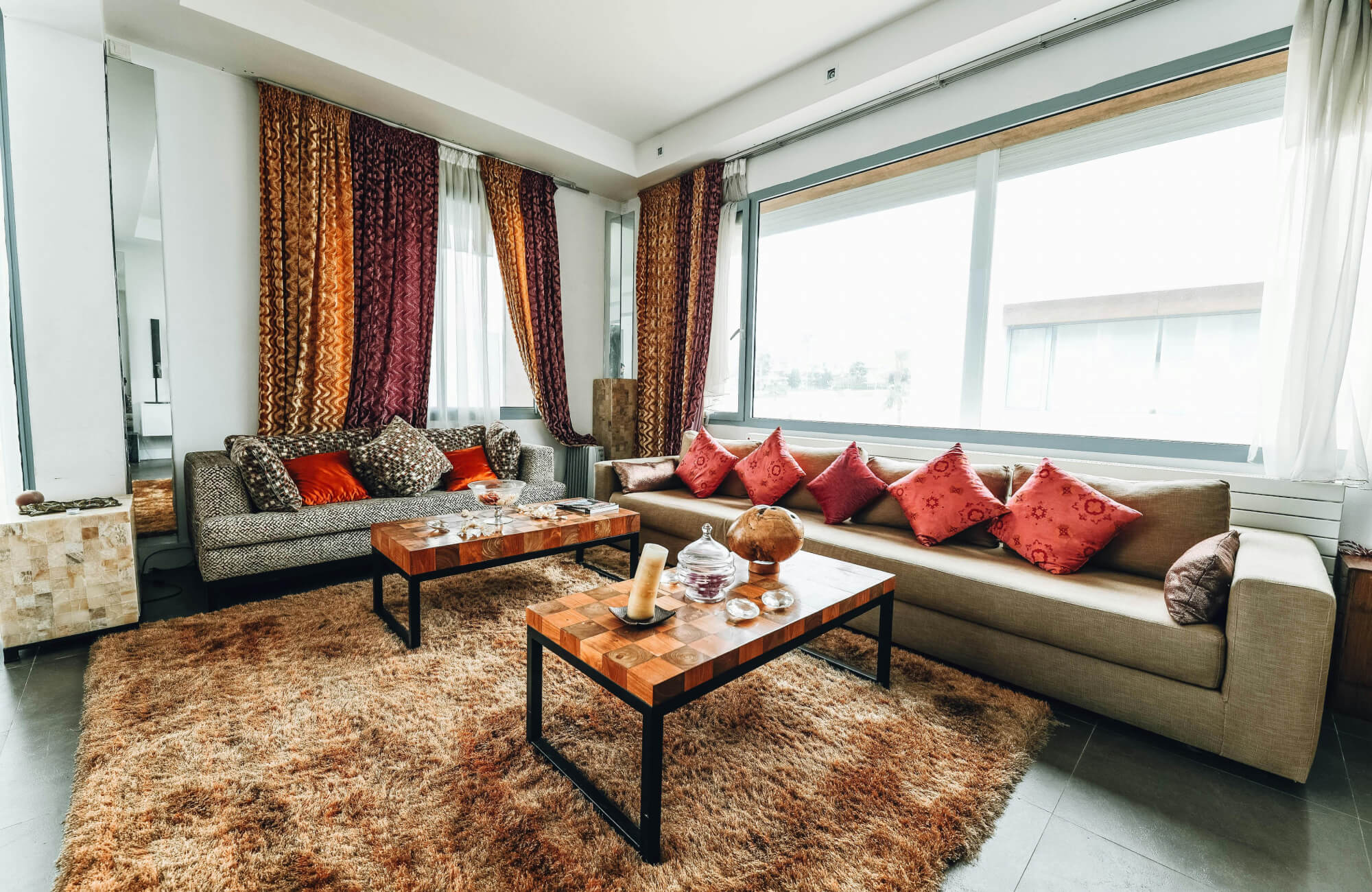 Contemporary bedroom with soft neutral seating, textured decorative pillows in rust, tan, and cream tones, and a sunlit window backdrop.
