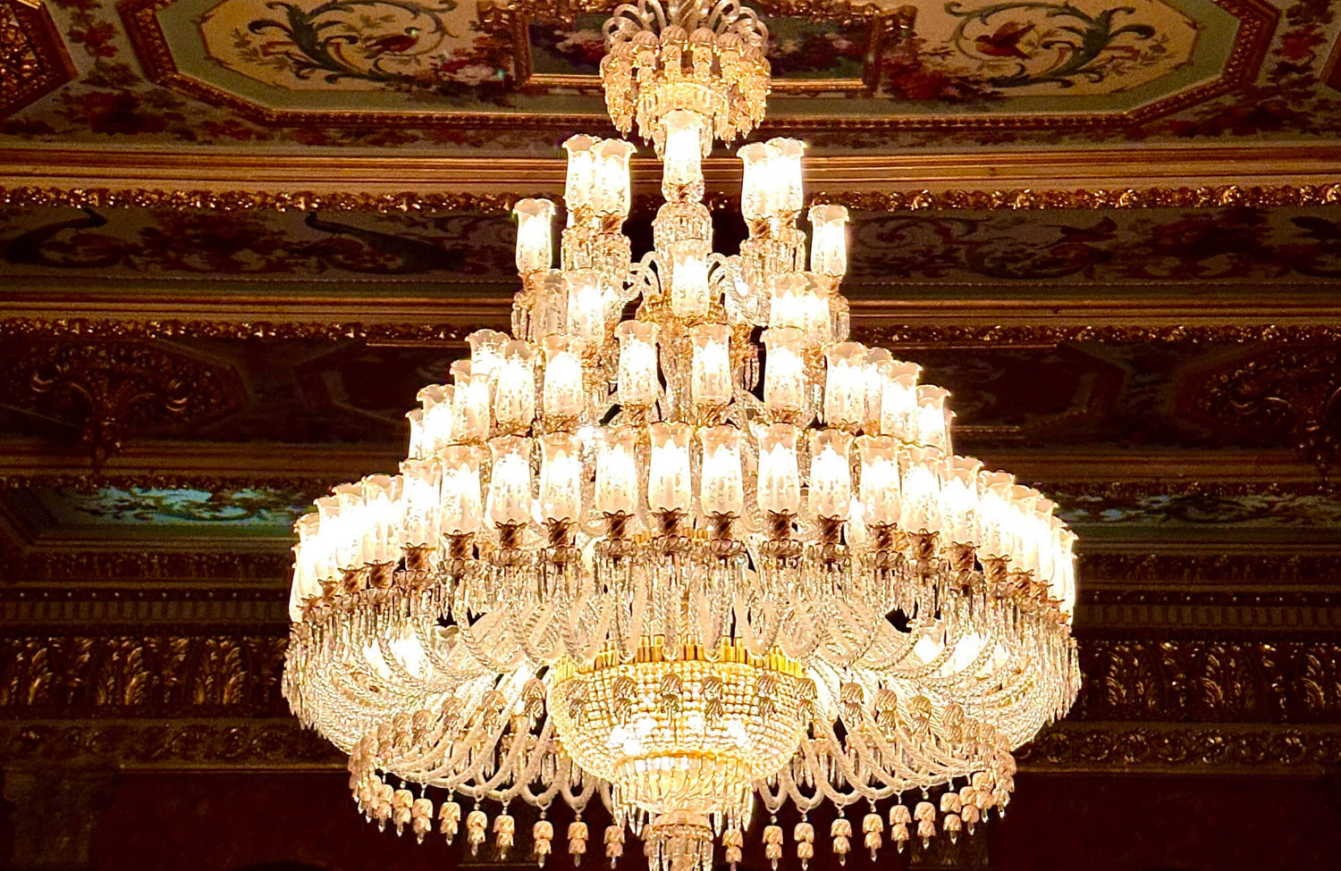 Opulent crystal chandelier with cascading tiers of glowing lights, creating a majestic focal point against the ornate, gilded ceiling.